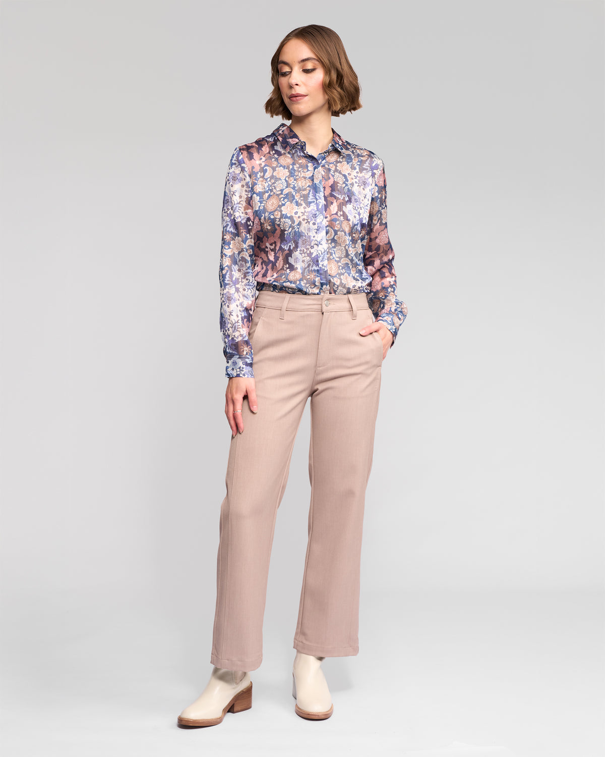 A woman with short, wavy hair stands against a light gray background wearing Vassalli's Neve Pant in Oyster, a beige straight-leg ankle-grazer with a tailored fit. Her left hand is in her pocket.