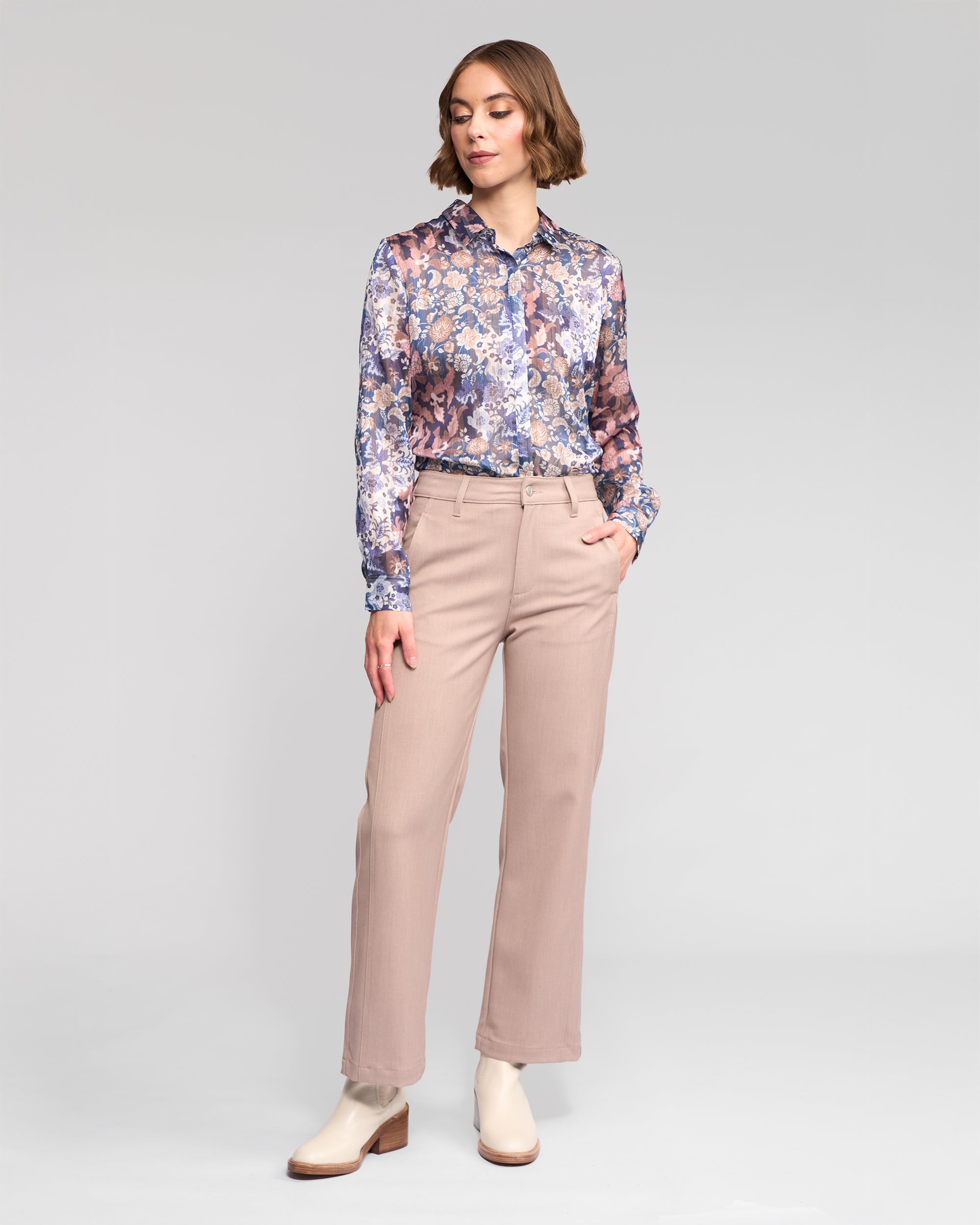 A woman with short, wavy hair stands against a light gray background wearing Vassalli's Neve Pant in Oyster, a beige straight-leg ankle-grazer with a tailored fit. Her left hand is in her pocket.