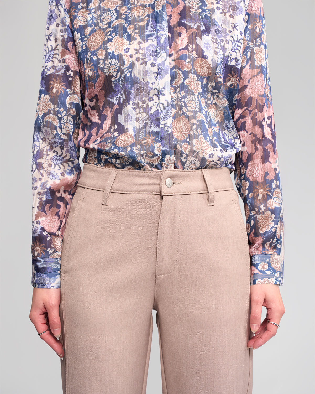 A person wears the Vassalli Neve Pant - Oyster, a high-waisted, straight-leg ankle-grazer, paired with a long-sleeved floral blouse, shown from shoulders to thighs against a plain background.