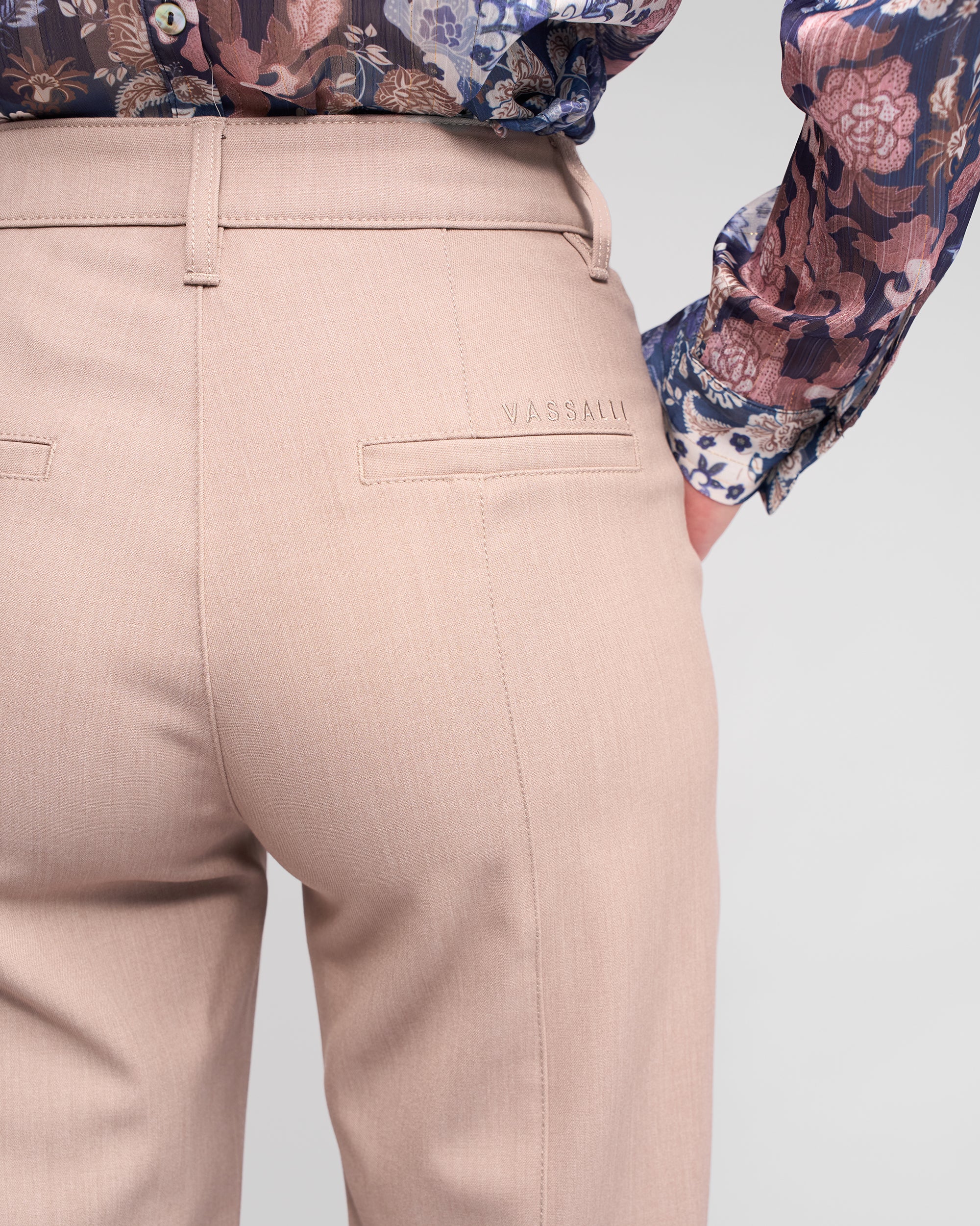 A close-up of a person wearing Vassalli’s Neve Pant in Oyster, straight-leg ankle-grazer style with double seam detailing and “VASSALLI” embroidered above the back pocket, paired with a tucked-in floral blouse.