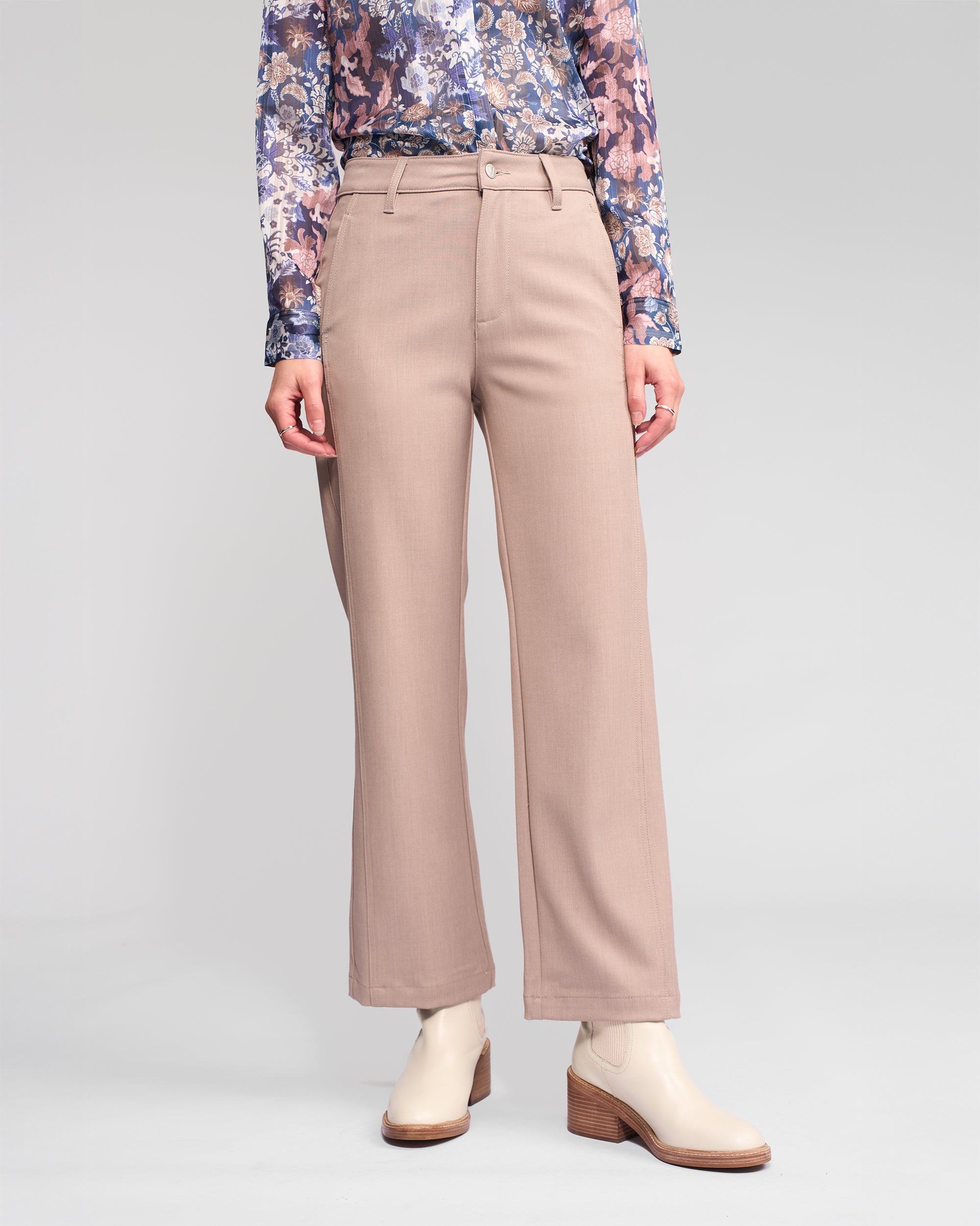 A person stands against a plain light background wearing Vassalli's Neve Pant in Oyster, styled with a floral long-sleeve top and cream ankle boots. Their face is not visible.