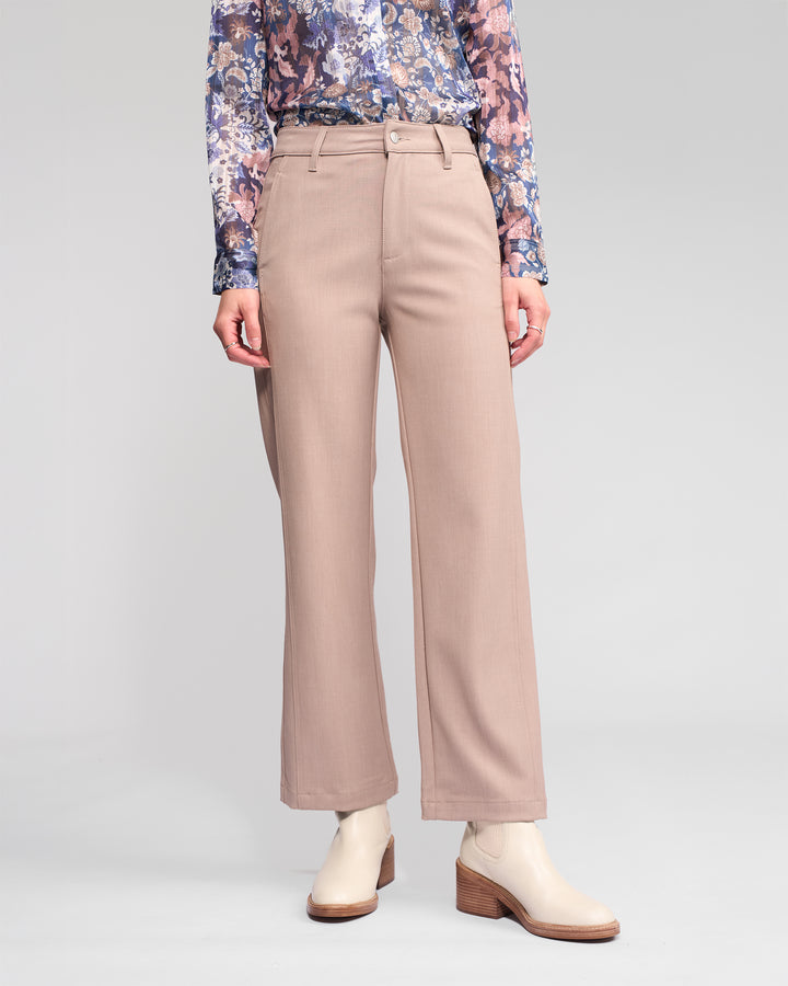A person stands against a plain light background wearing Vassalli's Neve Pant in Oyster, styled with a floral long-sleeve top and cream ankle boots. Their face is not visible.