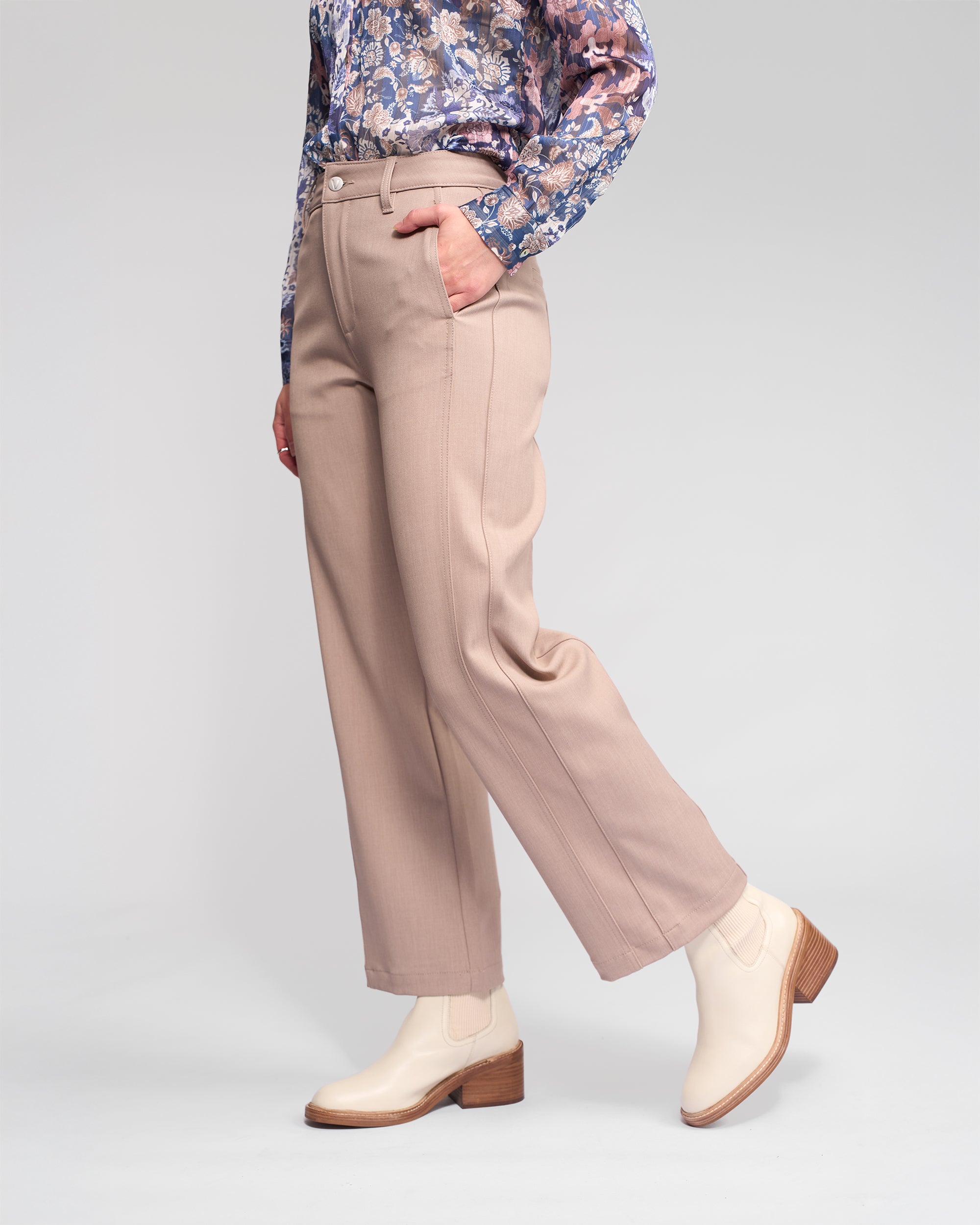 Someone stands against a neutral background wearing the Vassalli Neve Pant in Oyster, paired with a blue and purple floral long-sleeve blouse and cream ankle boots, one hand casually in their pocket.