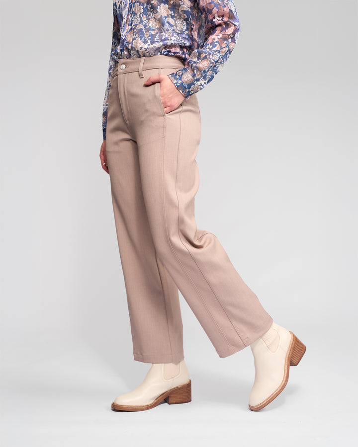 Someone stands against a neutral background wearing the Vassalli Neve Pant in Oyster, paired with a blue and purple floral long-sleeve blouse and cream ankle boots, one hand casually in their pocket.