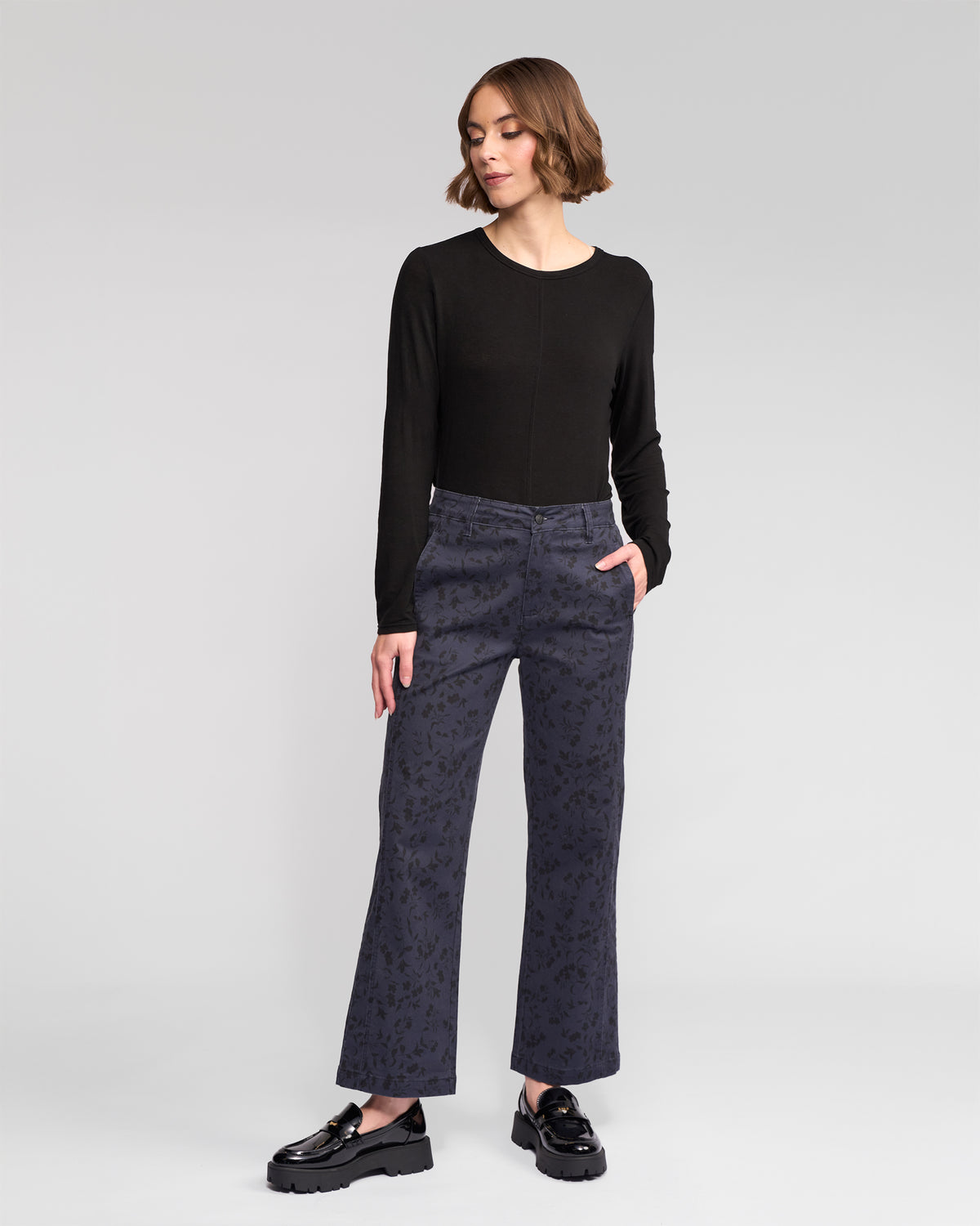 A woman with a short wavy hairstyle stands against a plain background, wearing a black long-sleeve top, Vassalli Neve Pant - Noir wide-leg denim, and black platform loafers, looking to the side with one hand in her pocket.