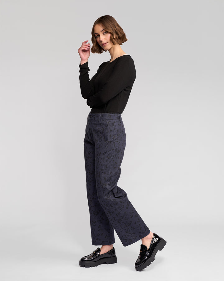 A woman with chin-length brown hair stands sideways, wearing a black long-sleeve top and Vassalli Neve Pant - Noir, paired with shiny black loafers, posing against a plain light gray background.