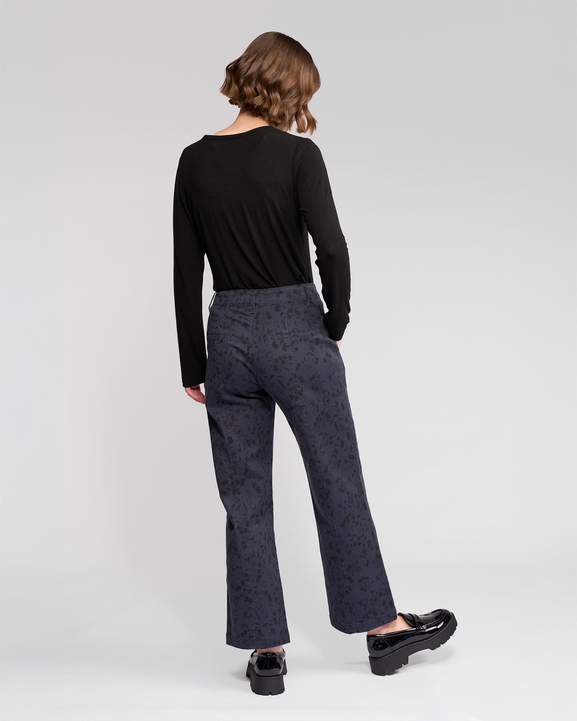 A person with short brown hair faces away, wearing a black long-sleeved top and Vassalli Neve Pant - Noir straight-leg ankle-grazer jeans with black platform shoes, against a plain light background.