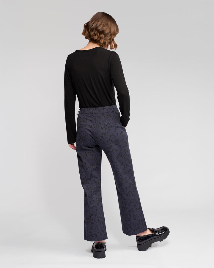 A person with short brown hair faces away, wearing a black long-sleeved top and Vassalli Neve Pant - Noir straight-leg ankle-grazer jeans with black platform shoes, against a plain light background.