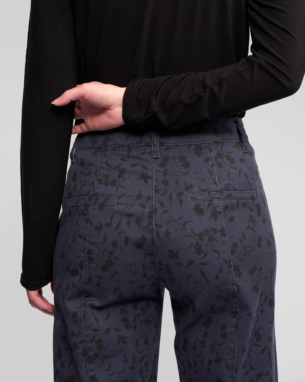 A person wears a long-sleeve black top and Vassalli's Neve Pant - Noir, featuring a subtle floral print and straight-leg ankle-grazer fit, seen from the back with one hand on their lower back for a polished denim look.