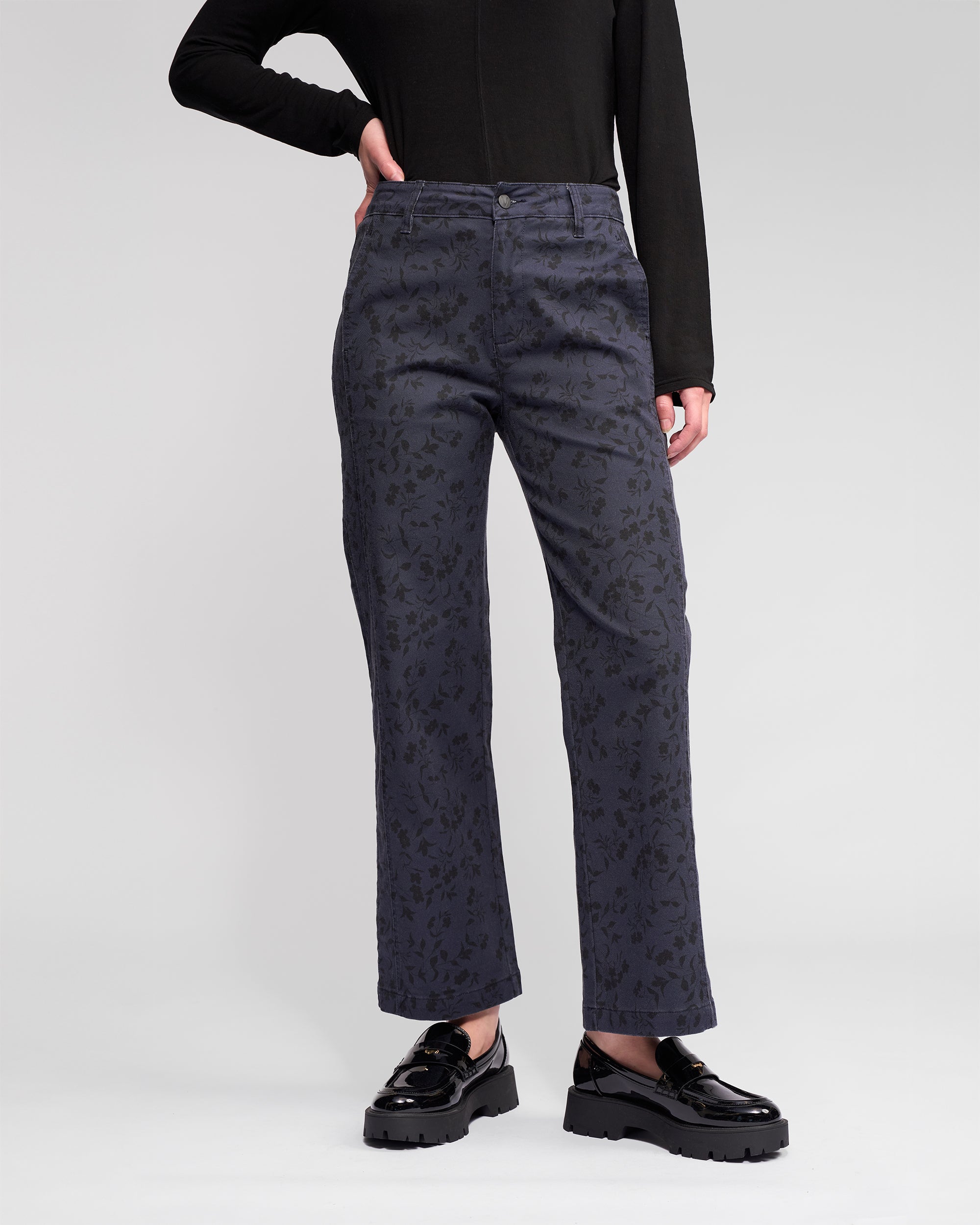 Person wears Vassalli’s Neve Pant – Noir, a high-waisted, straight-leg, ankle-grazer denim with a sleek edge, styled with black shiny platform shoes and a black long-sleeve top against a light background.