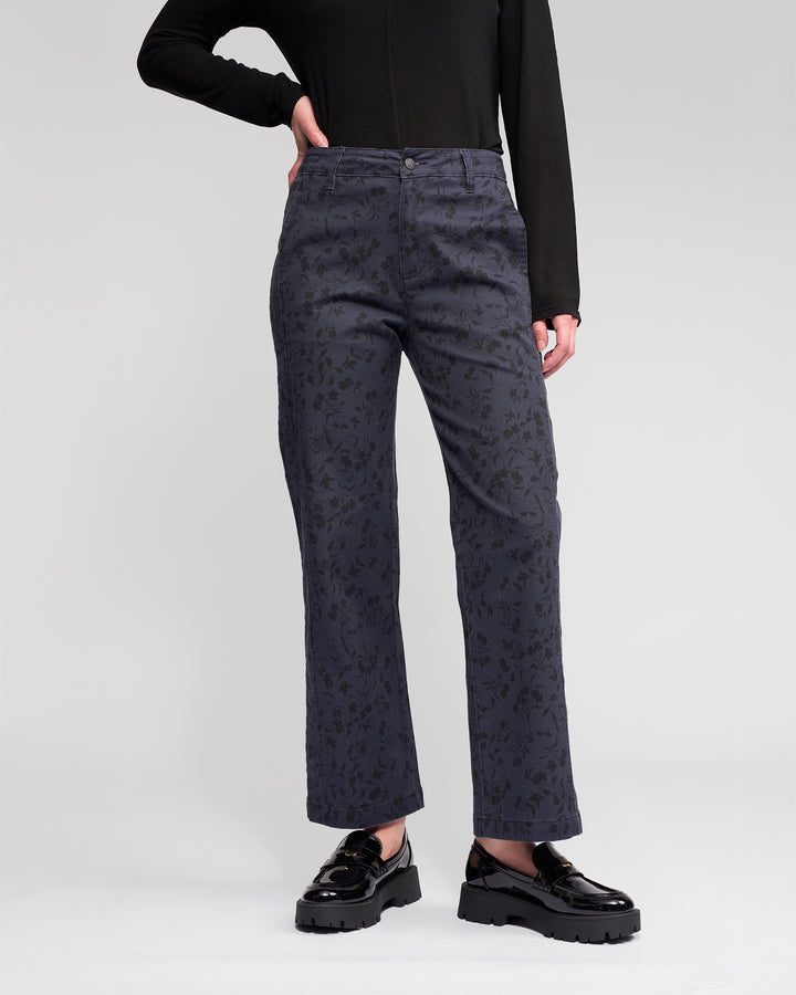 Person wears Vassalli’s Neve Pant – Noir, a high-waisted, straight-leg, ankle-grazer denim with a sleek edge, styled with black shiny platform shoes and a black long-sleeve top against a light background.