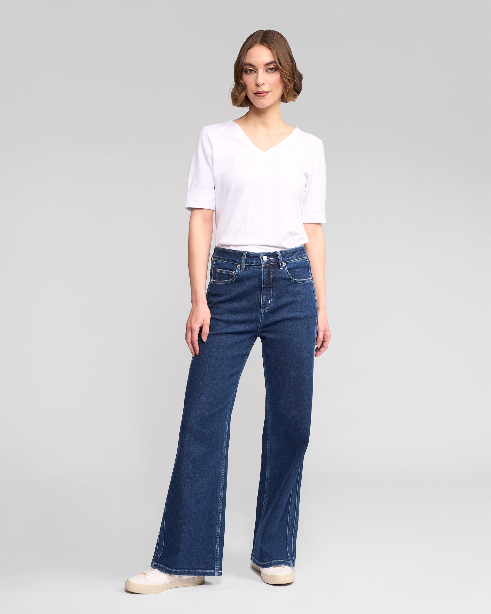A person with short, wavy brown hair and a neutral expression stands against a plain light gray background, wearing Vassalli's Wren Jean in Dark Blue, a white short-sleeve V-neck T-shirt, and white sneakers.