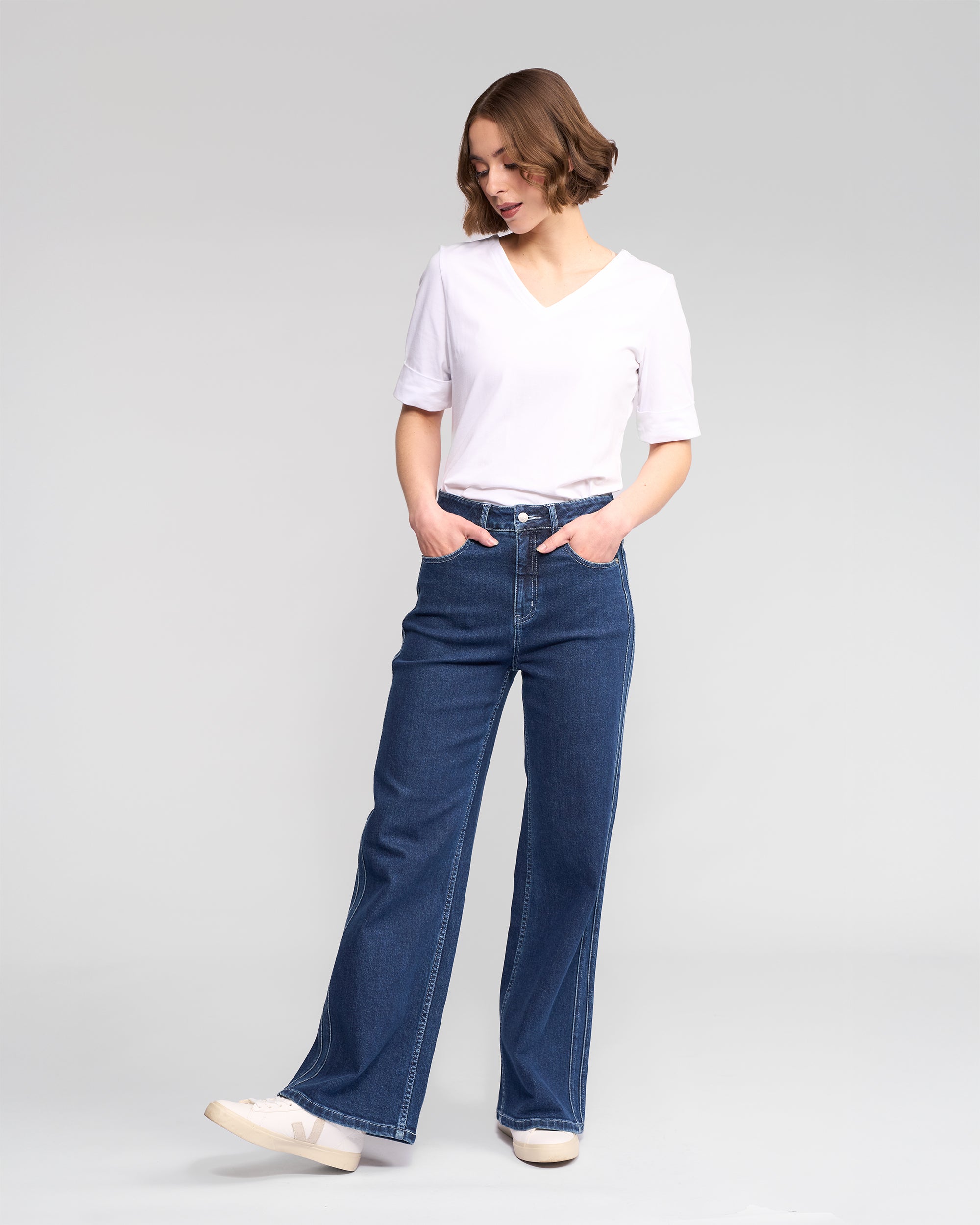 A person stands with hands in pockets, looking down, wearing a white V-neck top, Vassalli Wren Jean in dark blue, and white sneakers against a plain light gray background.