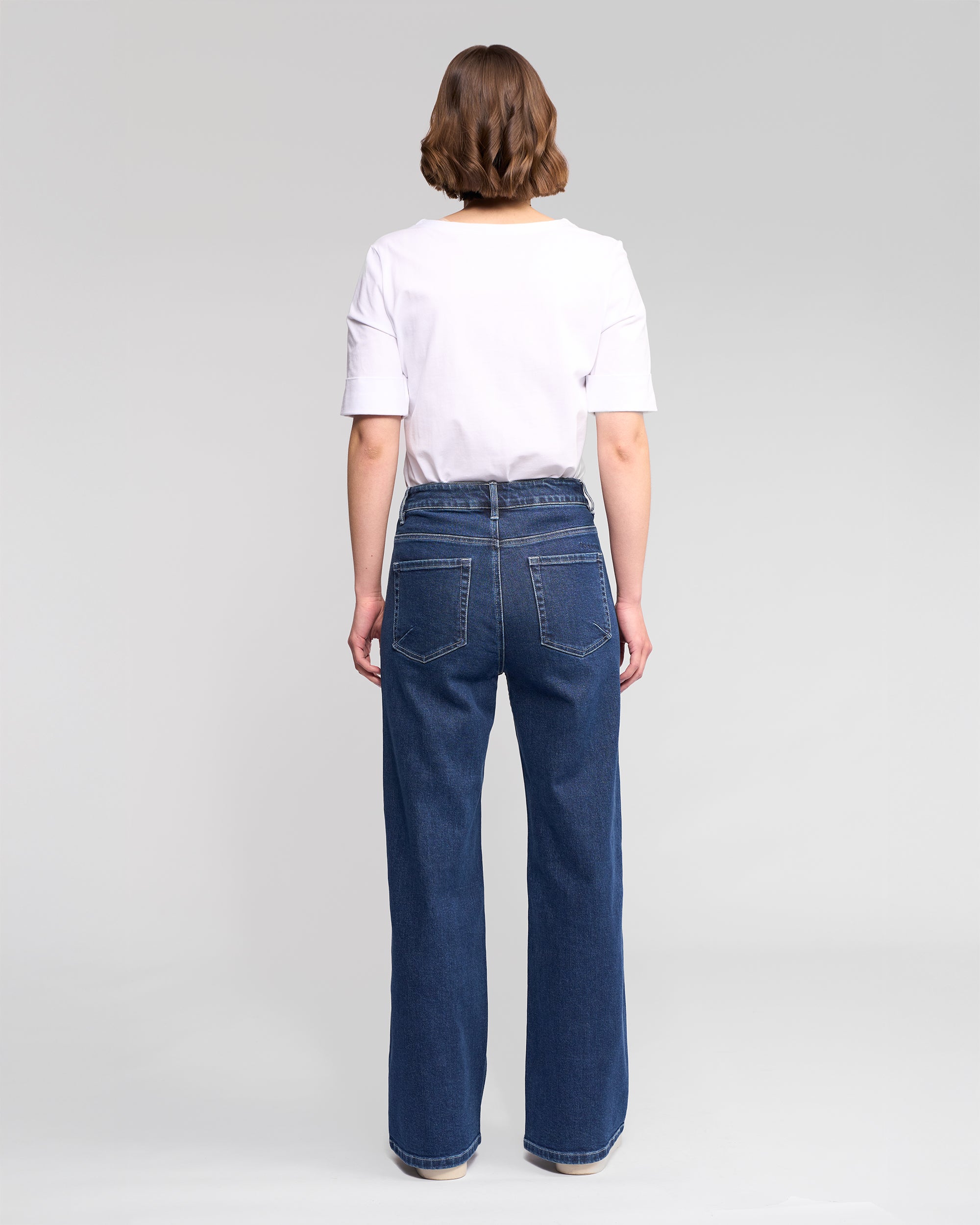 A person with short brown hair faces away, wearing a white T-shirt and Vassalli Wren Jean - Dark Blue, which are high-waisted, wide-leg jeans with back pockets, against a plain light gray background.