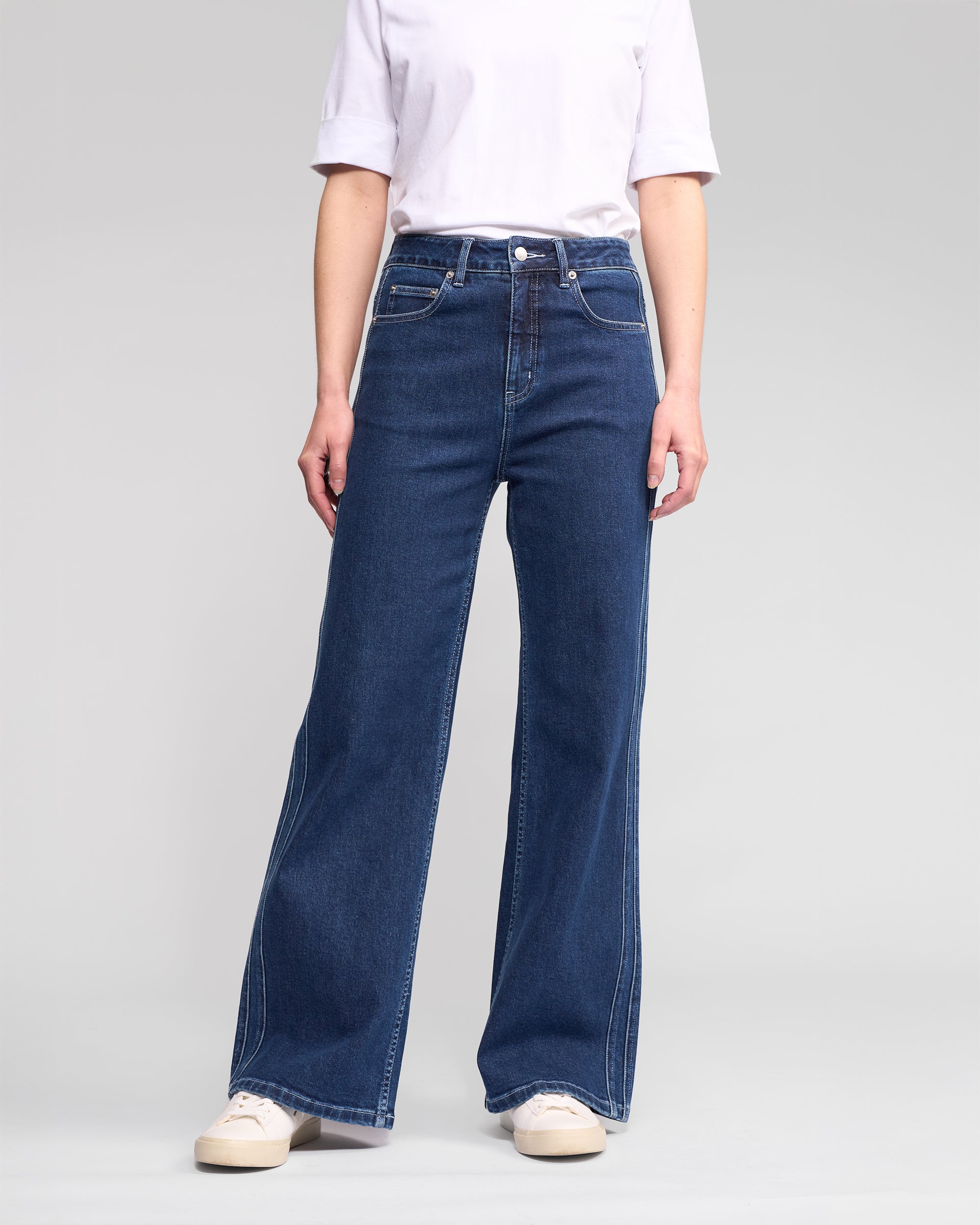 A person stands against a neutral background wearing the Vassalli Wren Jean in Dark Blue with a plain white t-shirt and white sneakers. Their face is not visible in the image.