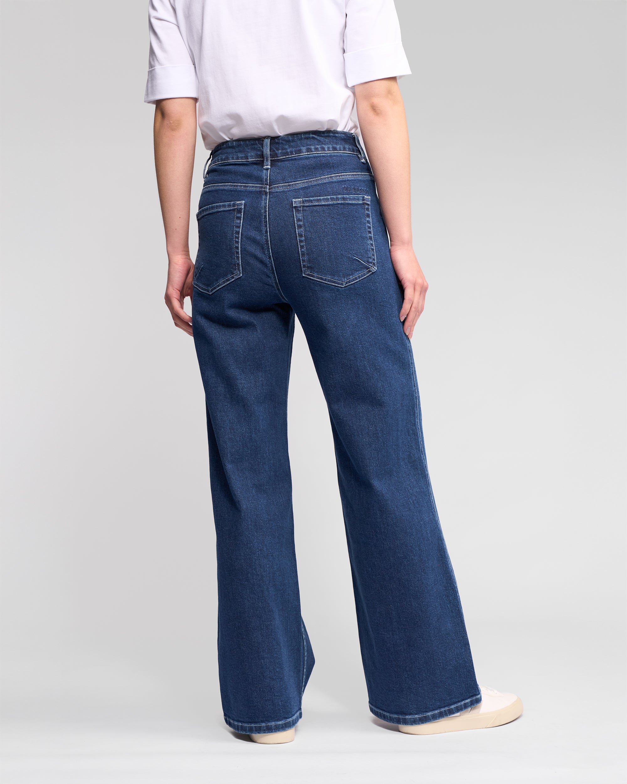 A person stands facing away, wearing a white short-sleeve shirt, Vassalli’s Wren Jean in dark blue—high-waisted, full-length, wide-leg—paired with white sneakers against a plain light background.