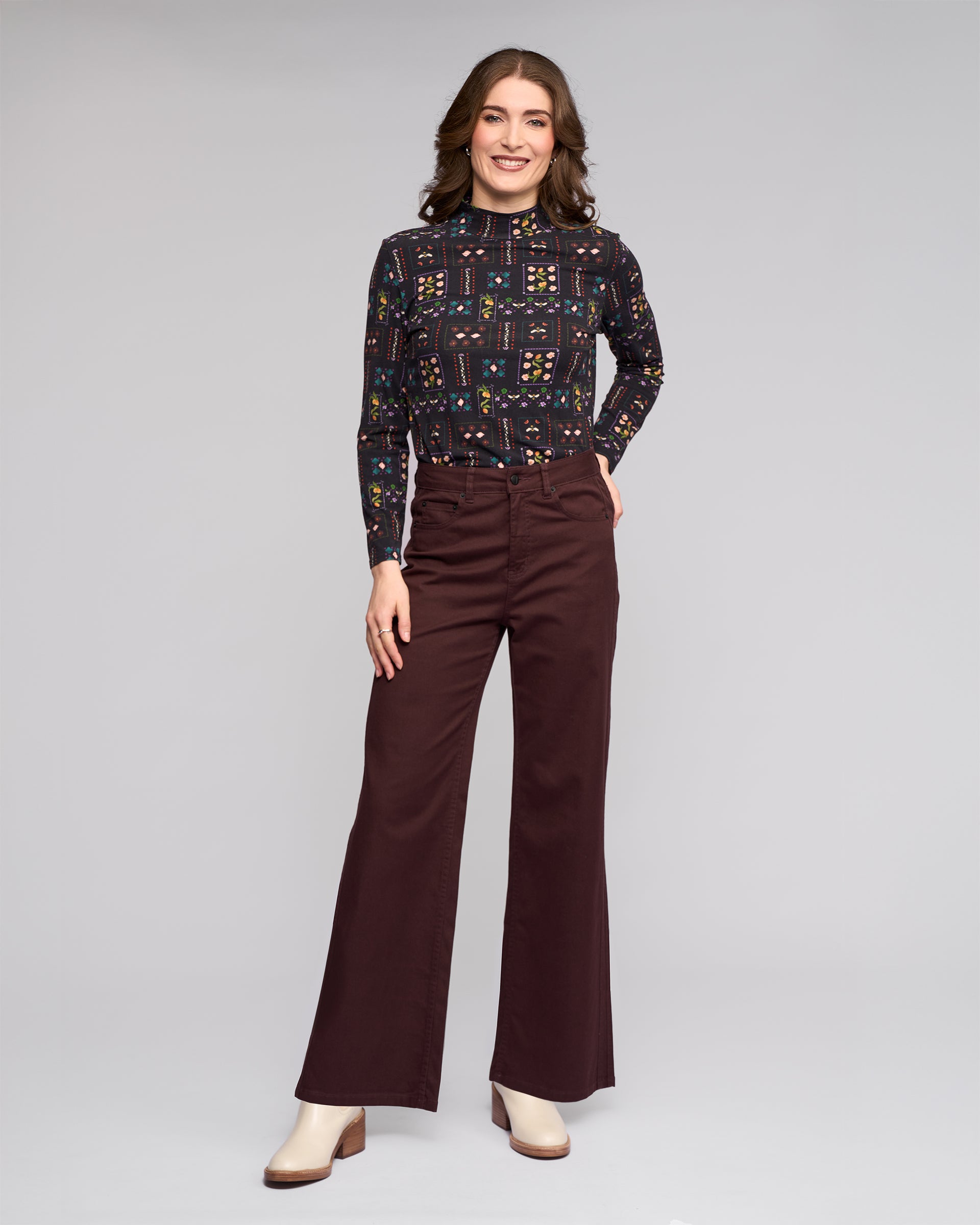 A woman smiles against a plain background, wearing the Vassalli Wren Jean in Merlot, paired with a long-sleeve patterned top and cream heeled boots.