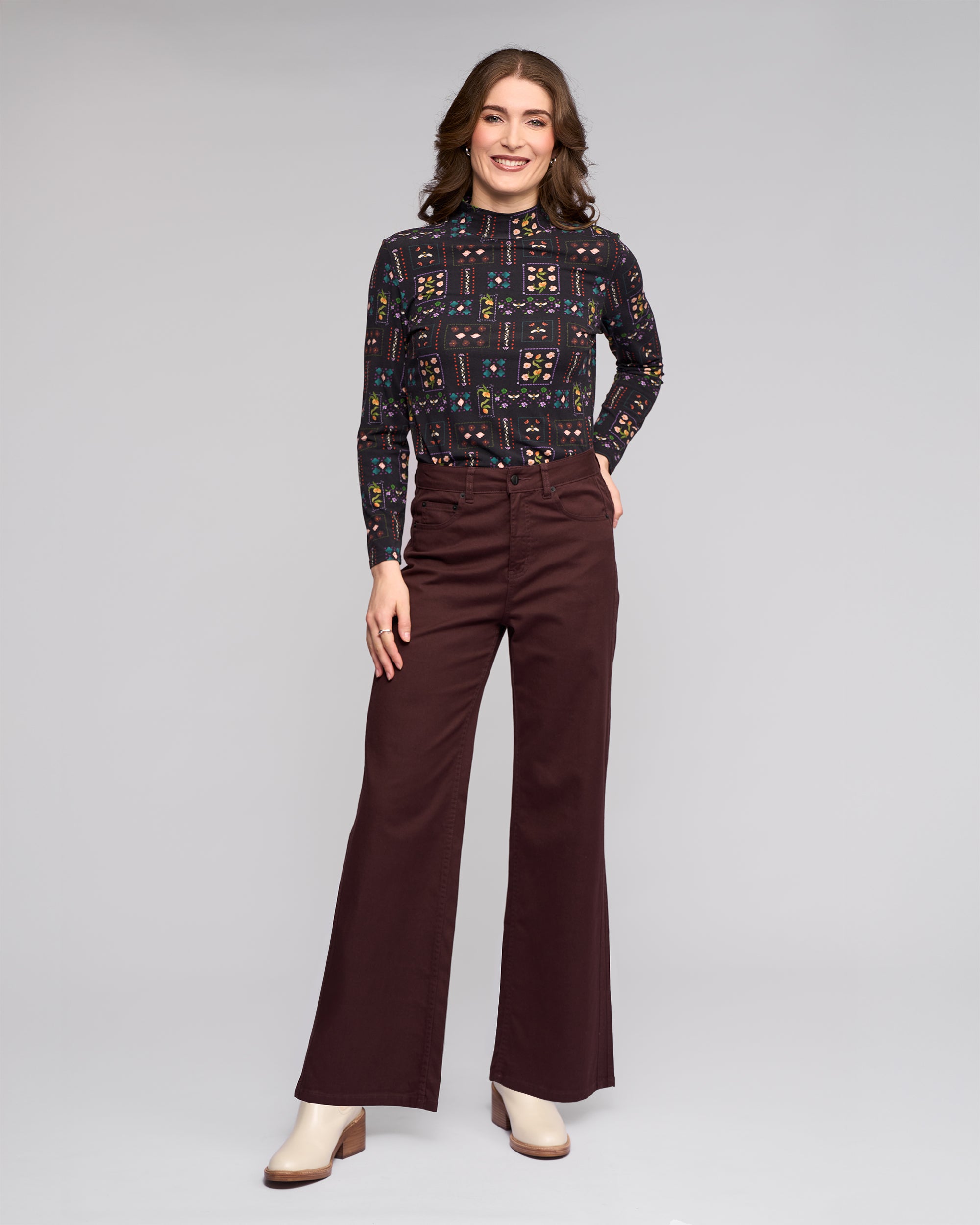 A woman smiles against a plain background, wearing the Vassalli Wren Jean in Merlot, paired with a long-sleeve patterned top and cream heeled boots.