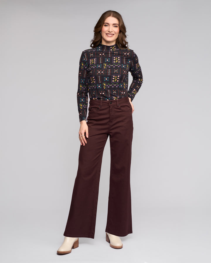 A woman smiles against a plain background, wearing the Vassalli Wren Jean in Merlot, paired with a long-sleeve patterned top and cream heeled boots.