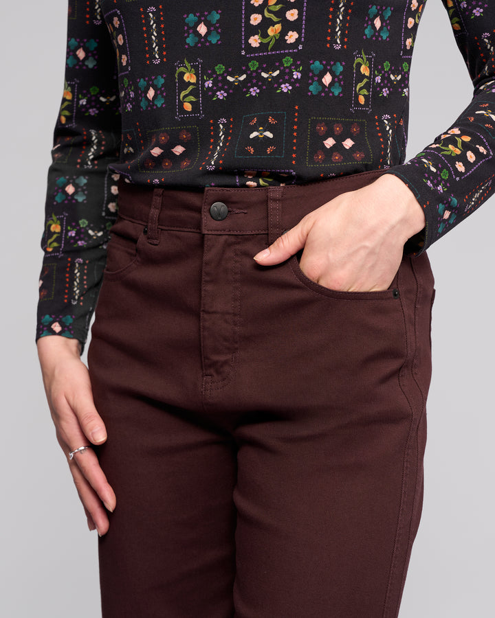 A person models the Vassalli Wren Jean - Merlot, styled with a long-sleeve patterned shirt featuring floral and geometric designs, one hand in a front pocket, the other resting by their side.