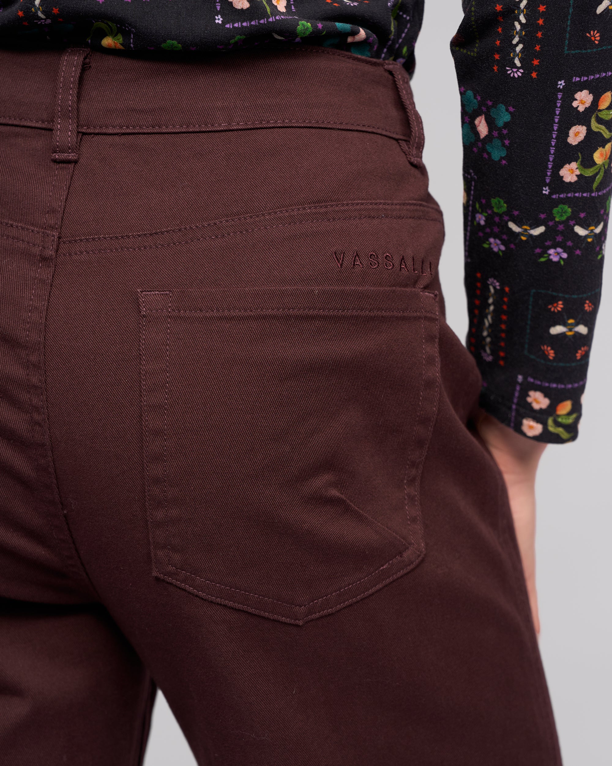 A close-up of a person wearing Vassalli's Wren Jean in Merlot with an embroidered label by the pocket, paired with a black floral long-sleeve top. Their hand rests at their side.