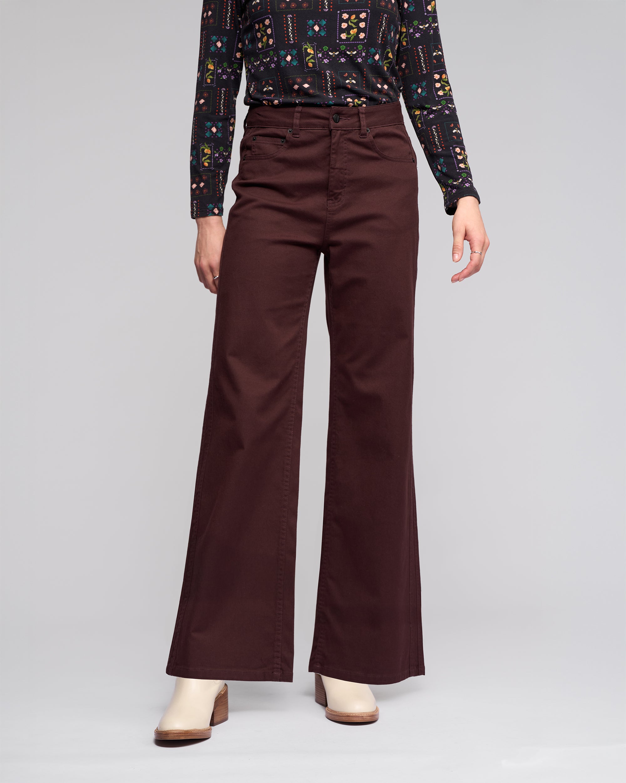 Model wears the Vassalli Wren Jean - Merlot, styled with a patterned long-sleeve shirt and off-white heeled shoes, standing against a plain light gray background. The upper part of the head is out of frame.