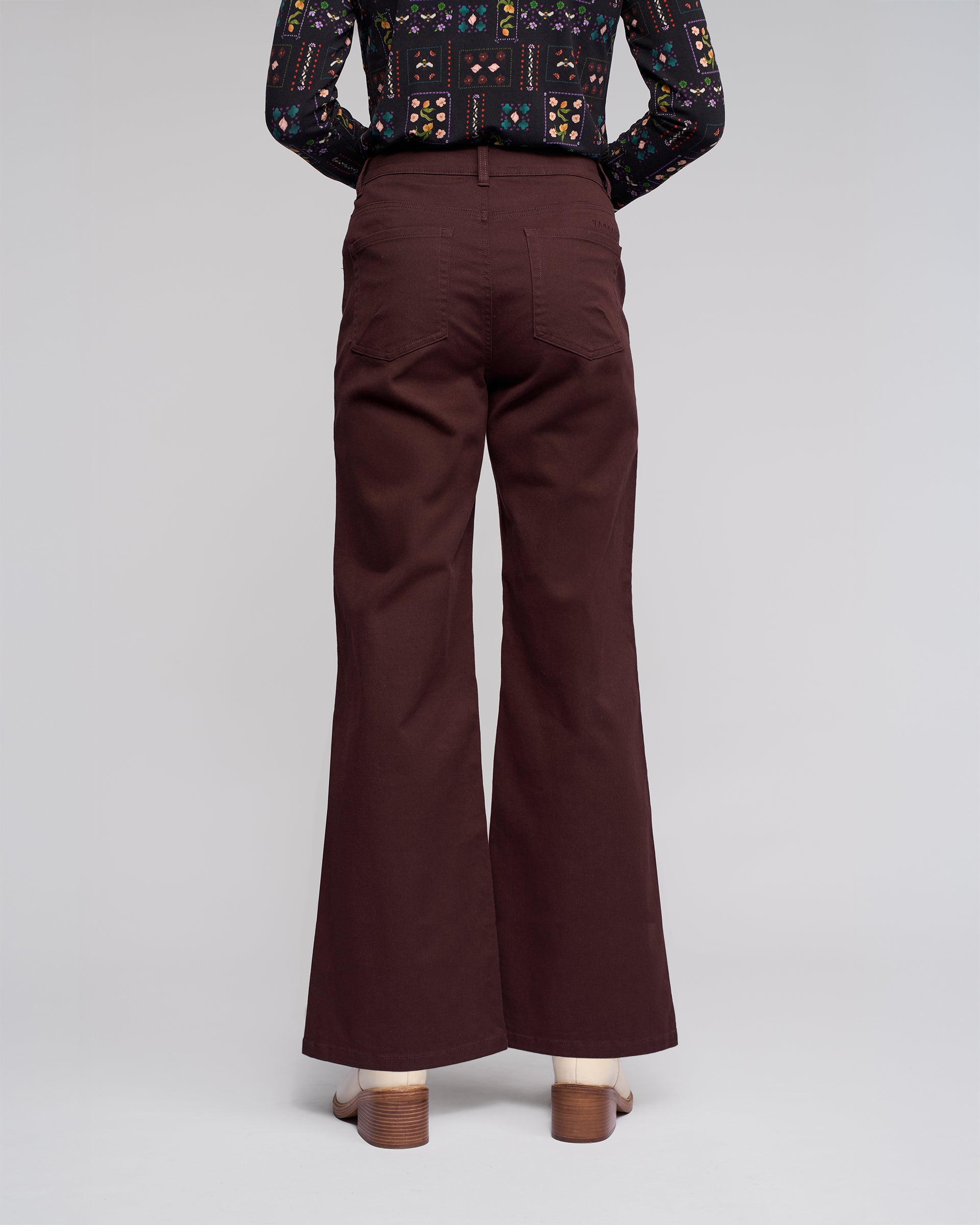 A person faces away, wearing Vassalli Wren Jean - Merlot wide-leg, high-waisted denim with back pockets and heeled sandals. A dark patterned long-sleeve shirt completes the look against a plain background.