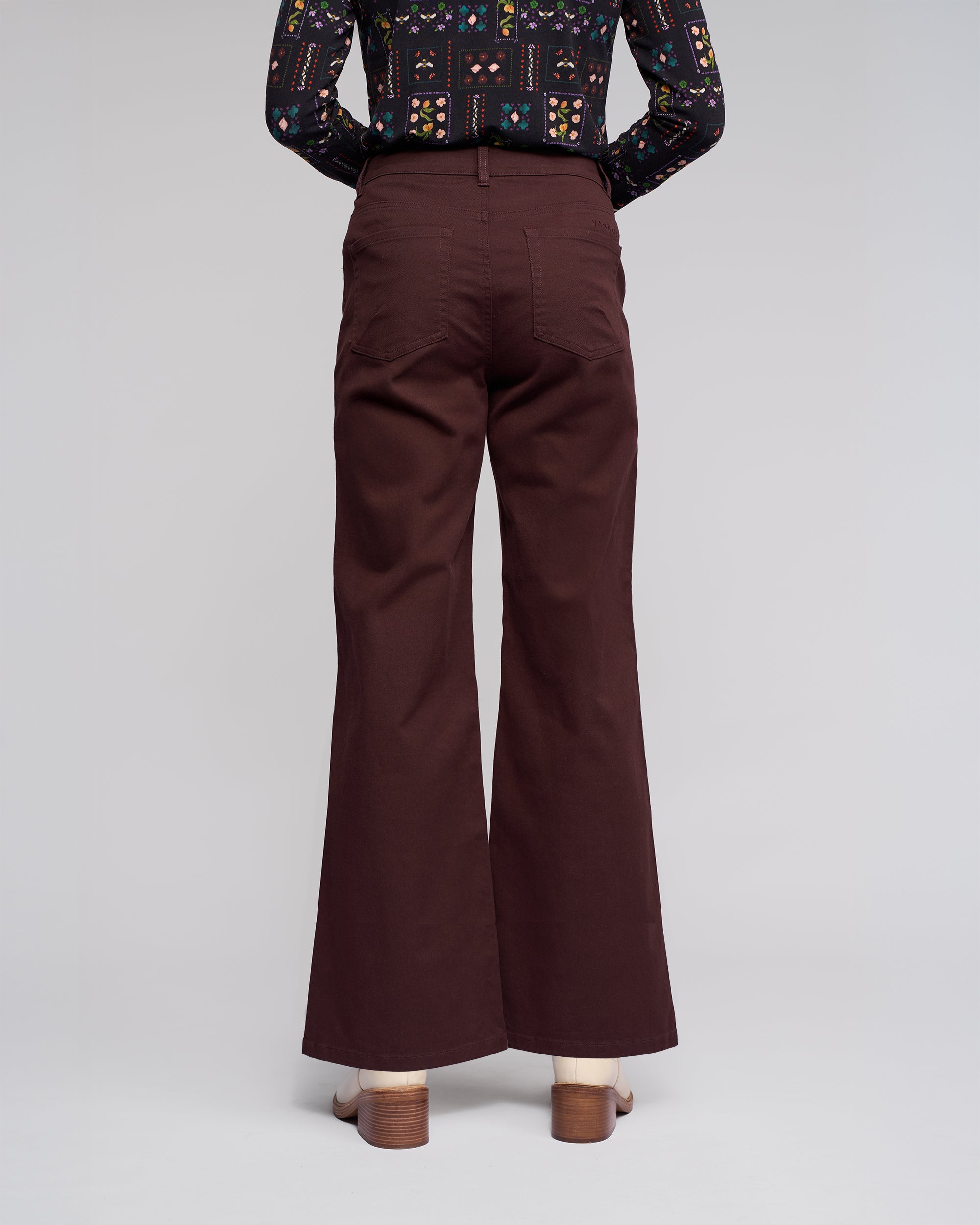 A person faces away, wearing Vassalli Wren Jean - Merlot wide-leg, high-waisted denim with back pockets and heeled sandals. A dark patterned long-sleeve shirt completes the look against a plain background.
