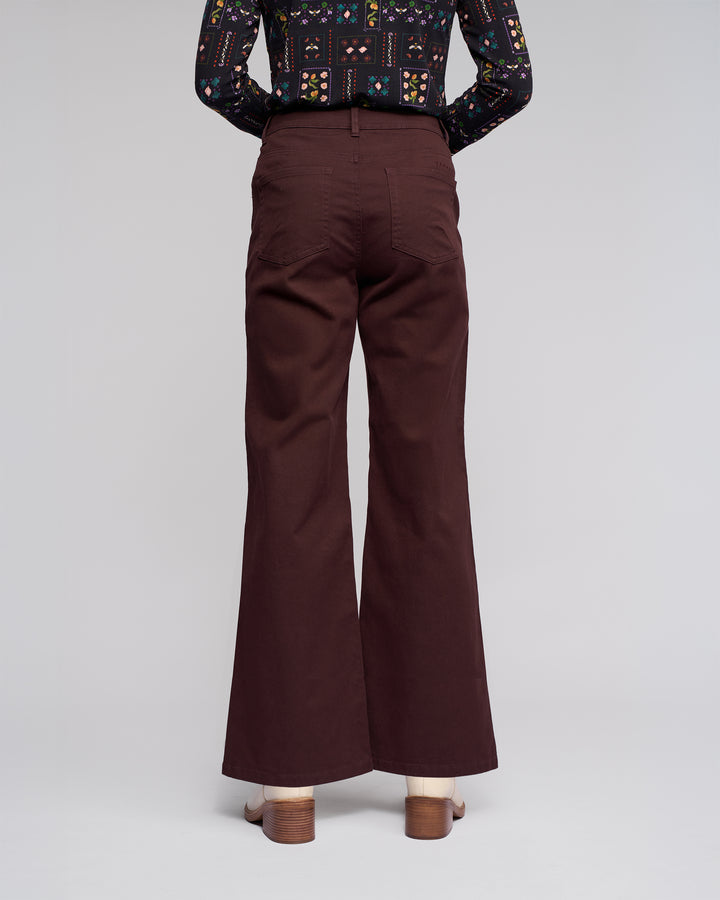 A person faces away, wearing Vassalli Wren Jean - Merlot wide-leg, high-waisted denim with back pockets and heeled sandals. A dark patterned long-sleeve shirt completes the look against a plain background.
