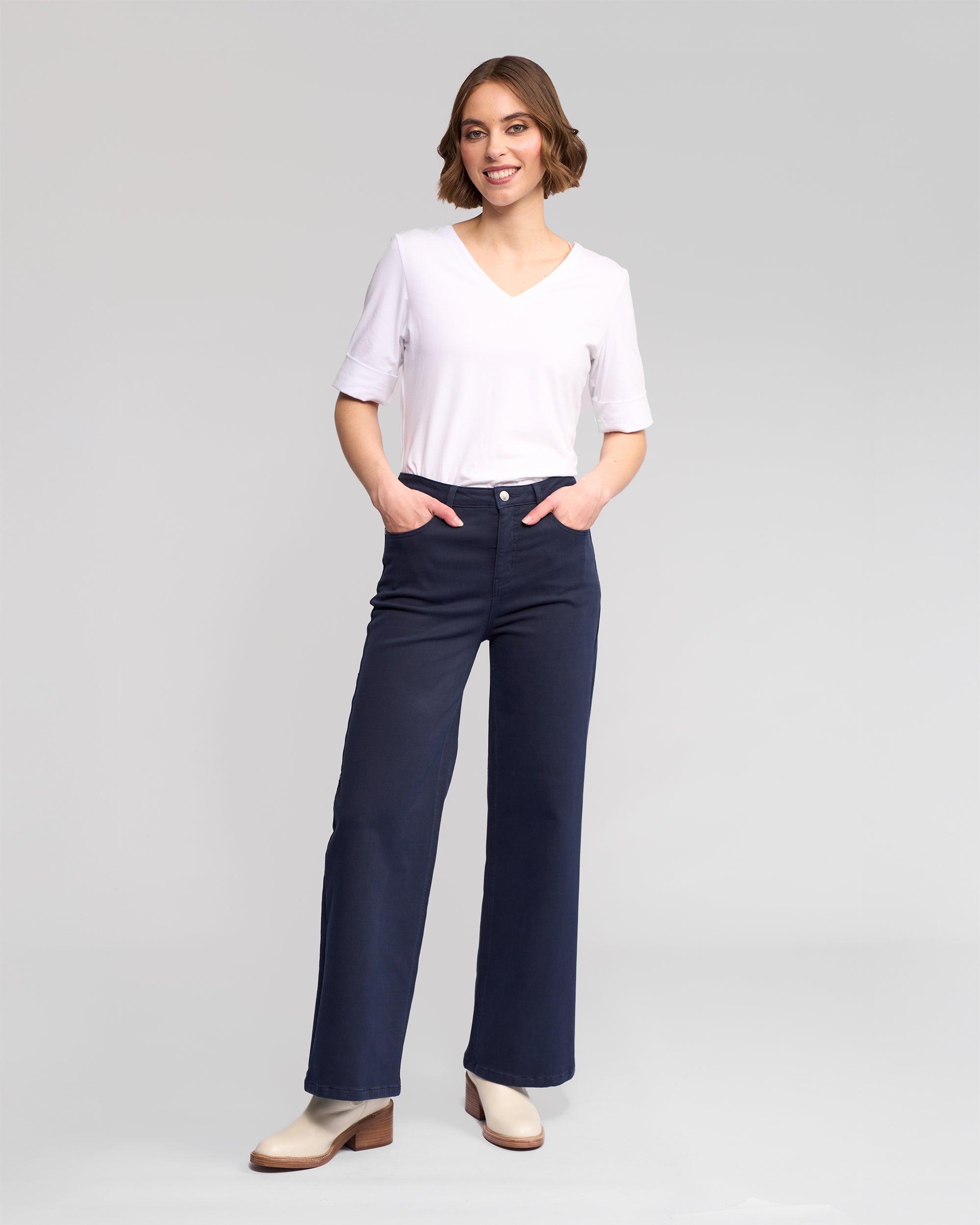 A woman smiles with hands in the pockets of Vassalli's Solo Pant - Carbon, a dark blue winter-weight wide-leg knit style. She pairs them with a white short-sleeve V-neck top and beige heeled boots, posing before a plain light gray backdrop.