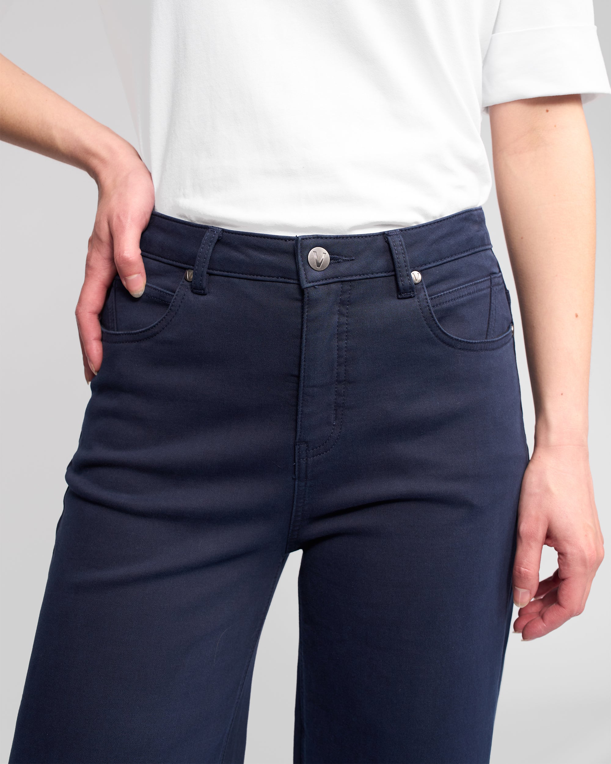A person models the Vassalli Solo Pant - Carbon, high-waisted and wide-leg in dark blue, paired with a white short-sleeve shirt. The image highlights the cotton-tencel stretch fabric at the waist and hips.