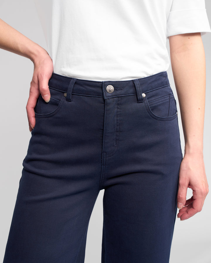A person models the Vassalli Solo Pant - Carbon, high-waisted and wide-leg in dark blue, paired with a white short-sleeve shirt. The image highlights the cotton-tencel stretch fabric at the waist and hips.