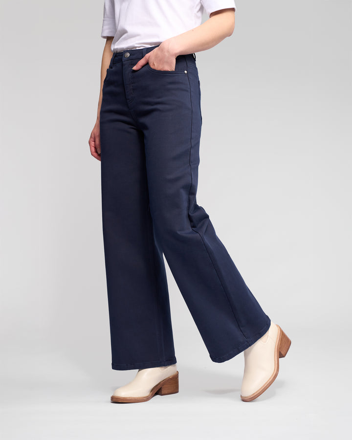 A person wears the Vassalli Solo Pant - Carbon, paired with a white shirt and white heeled ankle boots, standing with one hand in their pocket against a plain light gray background. Their face is not visible.
