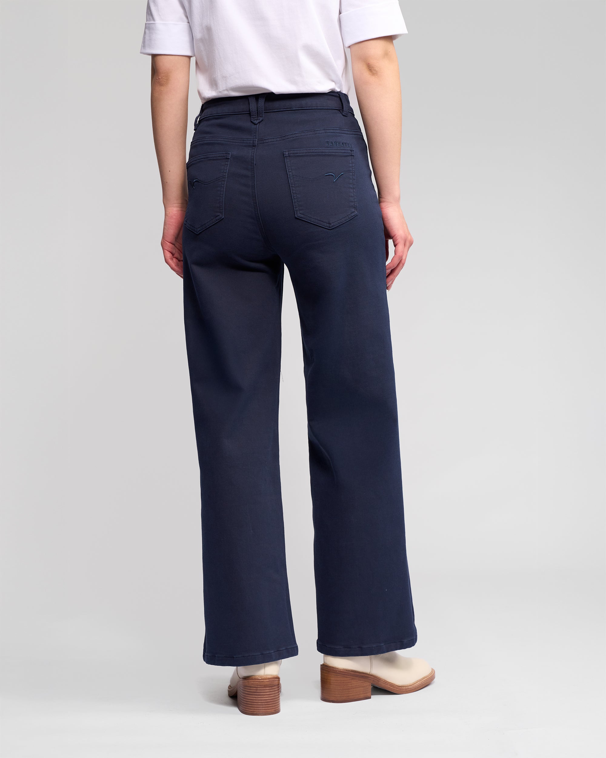 A person faces away, wearing a white short-sleeve shirt and Vassalli's Solo Pant - Carbon, high-waisted wide-leg jeans in dark blue knit, paired with beige heeled shoes against a plain light gray background.