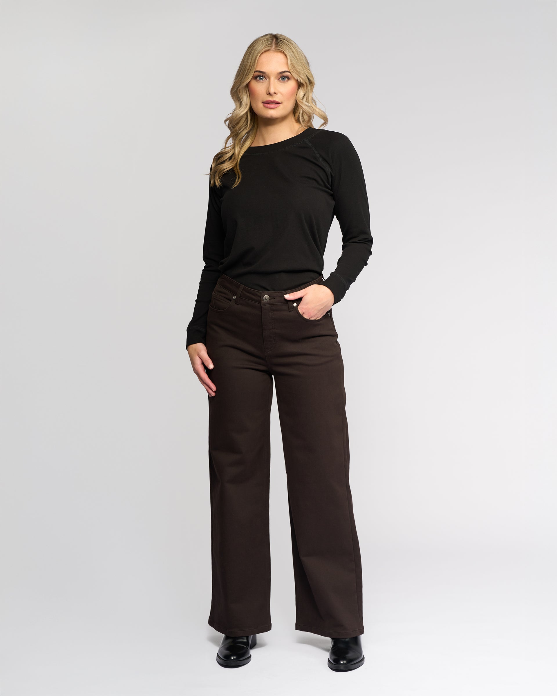A woman with long blonde hair stands against a plain light background, wearing the Vassalli Solo Pant in Chocolate with a black long-sleeve top and black shoes. One hand rests in her pocket as she looks slightly to the side.