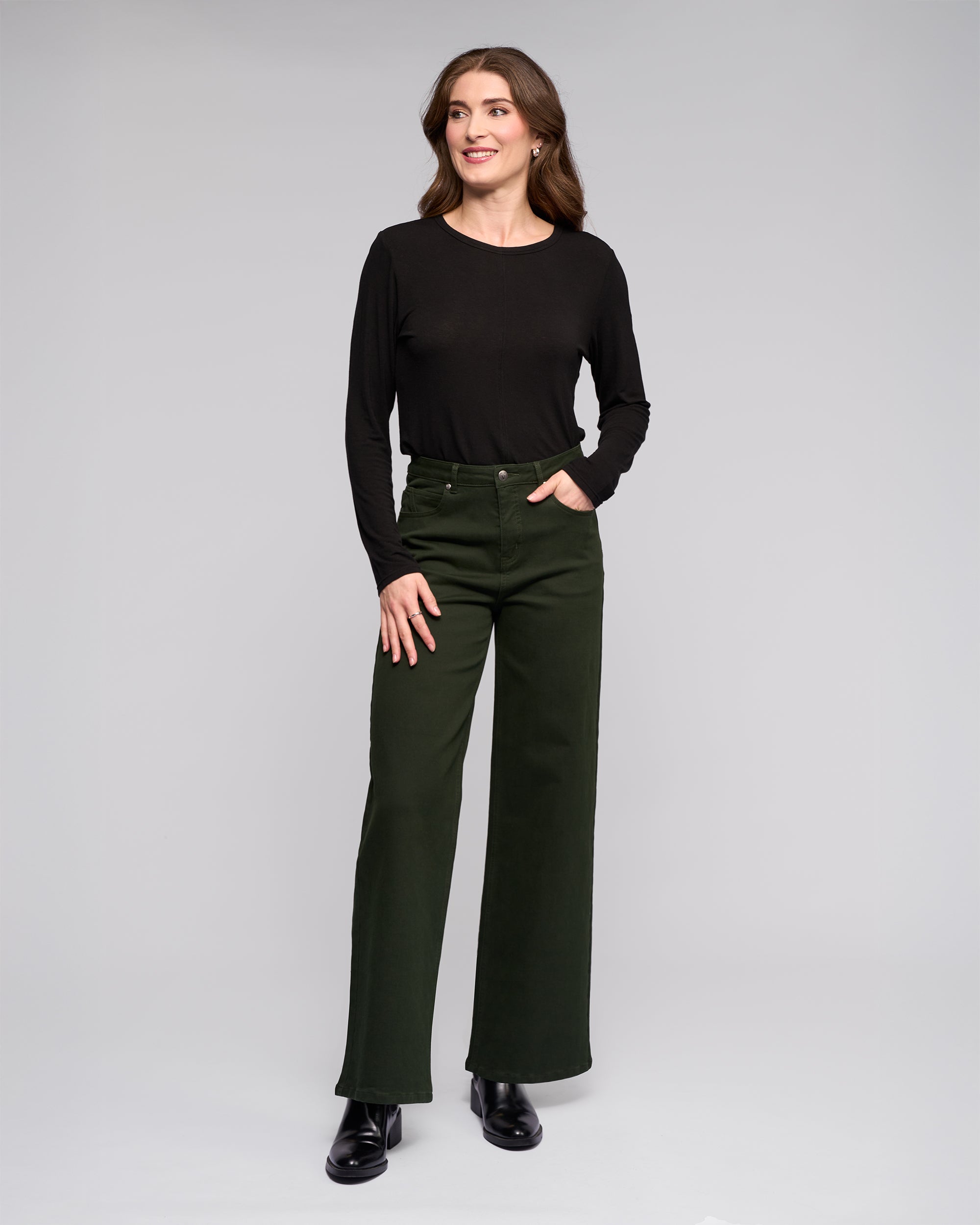 A woman with long brown hair smiles while looking to the side, wearing Vassalli’s Solo Pant in Forest and a black long-sleeve top, standing against a light grey background with black boots.