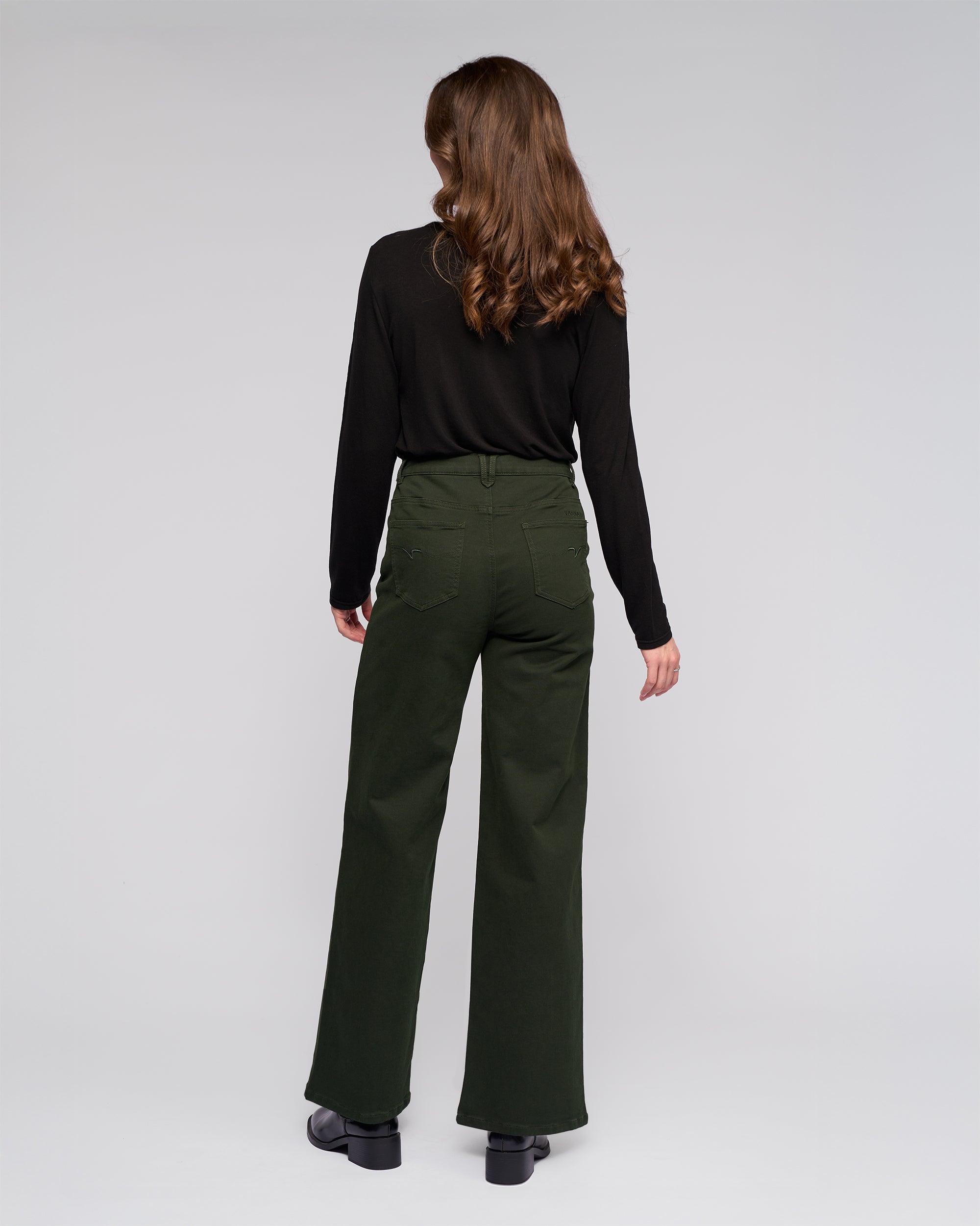 A woman with long brown hair stands facing away, wearing a black long-sleeve top, Vassalli's Solo Pant - Forest, and black shoes against a plain light background.