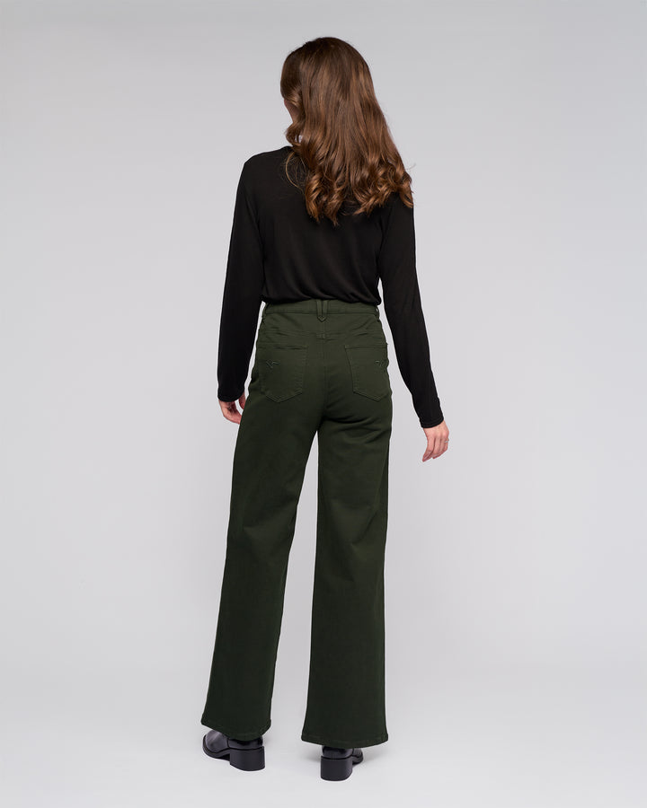 A woman with long brown hair stands facing away, wearing a black long-sleeve top, Vassalli's Solo Pant - Forest, and black shoes against a plain light background.
