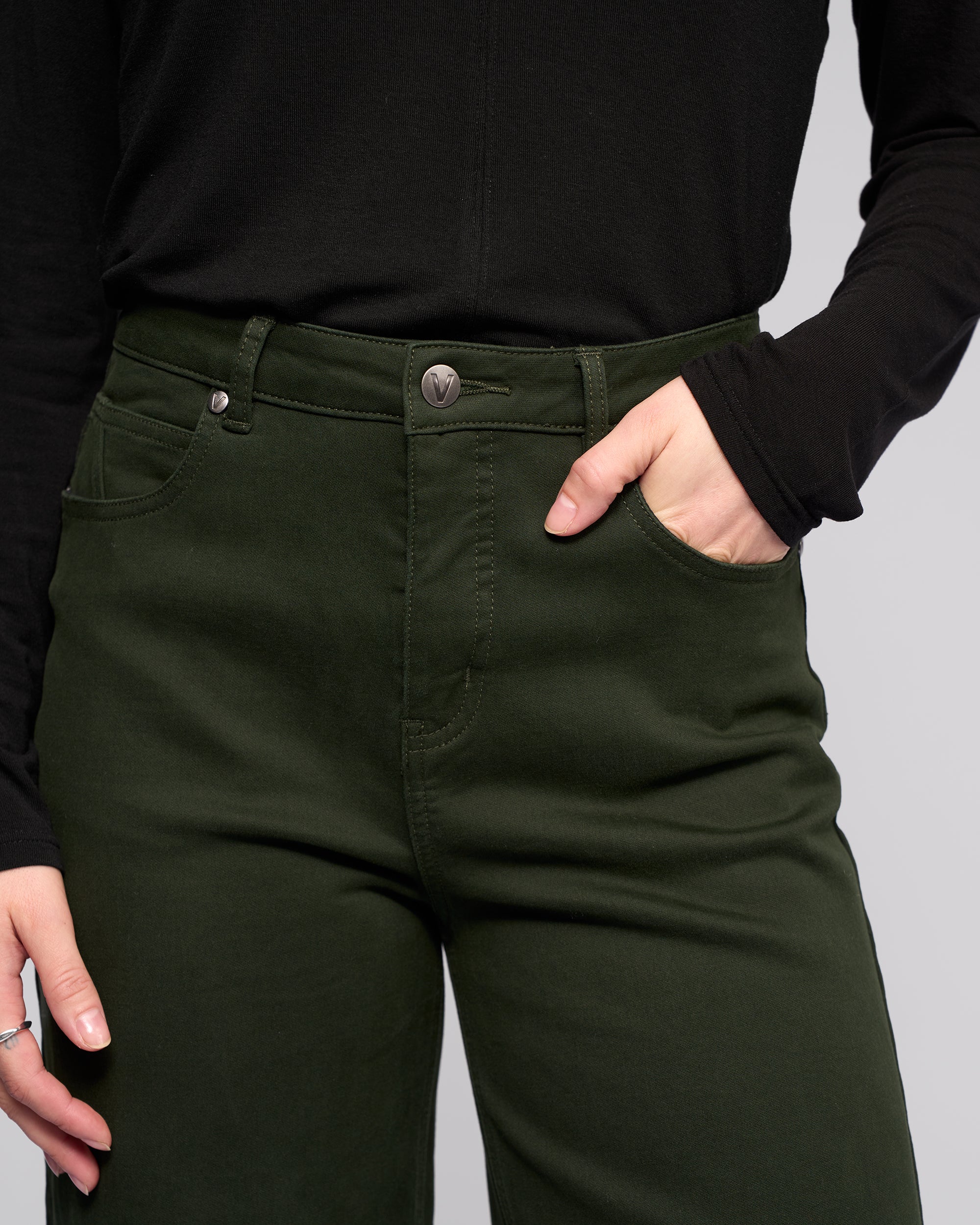 A person wearing the Vassalli Solo Pant - Forest, a pair of high-waisted, wide-leg knit jeans, and a black long-sleeve shirt stands with one hand in their pocket against a plain light gray background.
