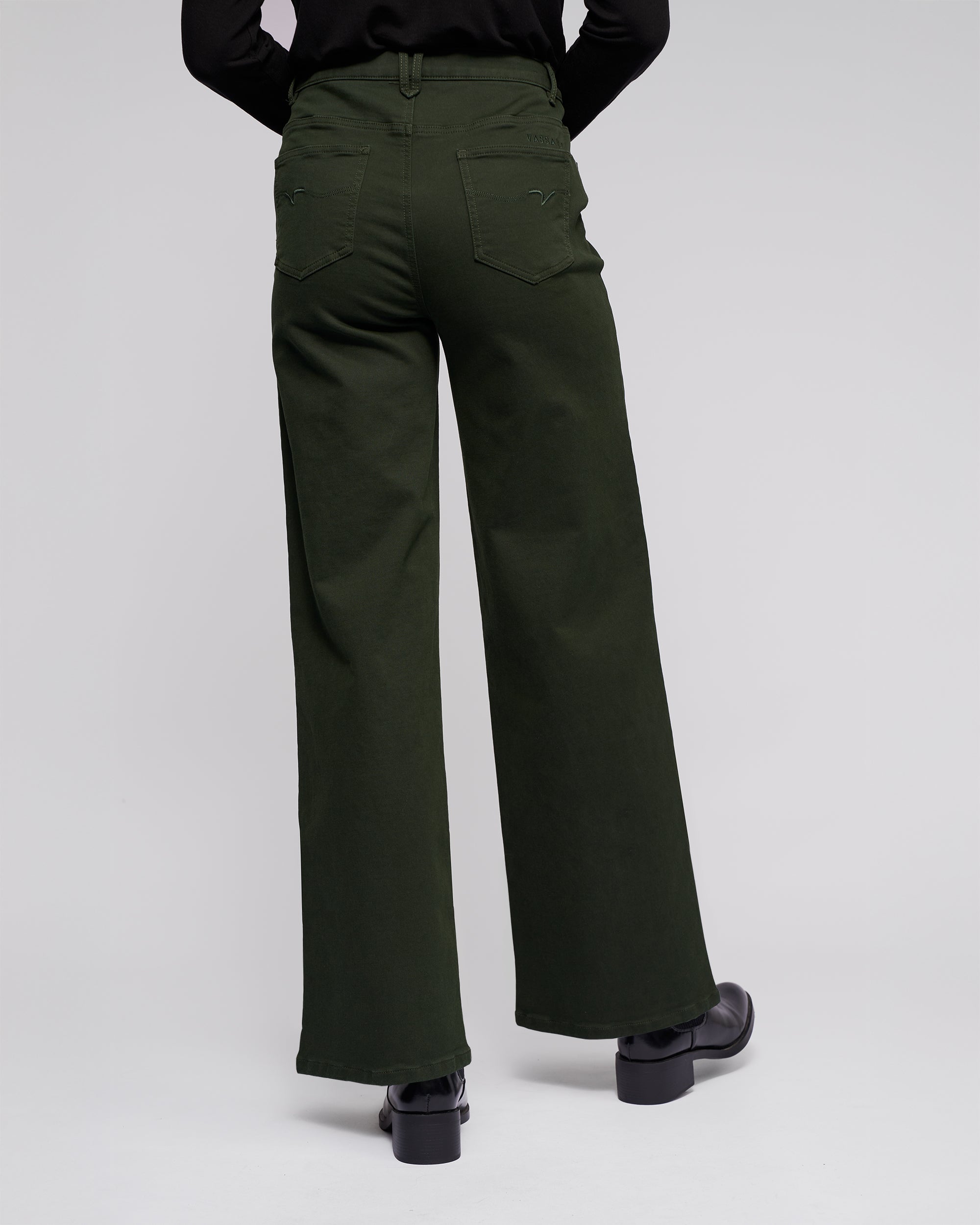 A person wearing the Vassalli Solo Pant - Forest, a high-waisted wide-leg knit jean in forest green cotton tencel, paired with black shoes, stands with their back to the camera against a plain light background.