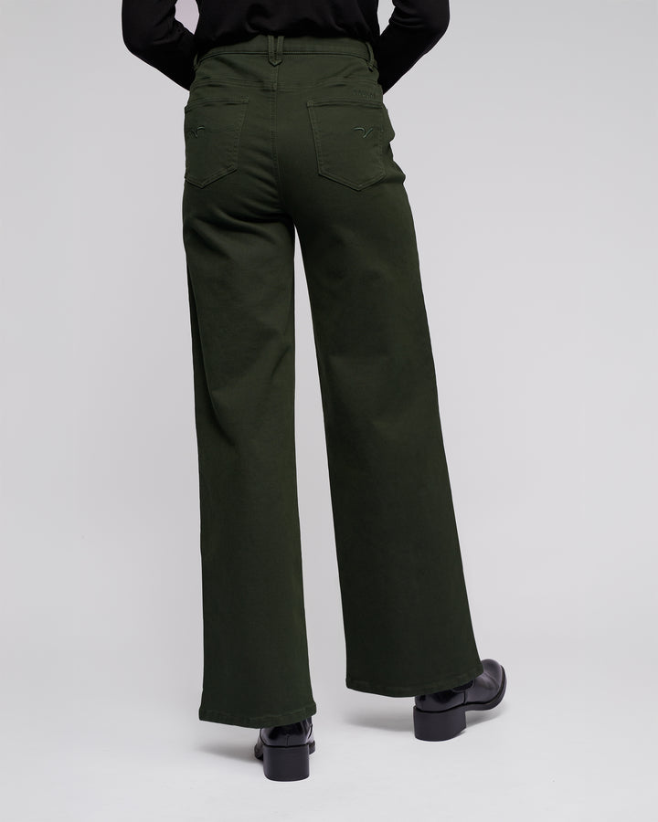 A person wearing the Vassalli Solo Pant - Forest, a high-waisted wide-leg knit jean in forest green cotton tencel, paired with black shoes, stands with their back to the camera against a plain light background.