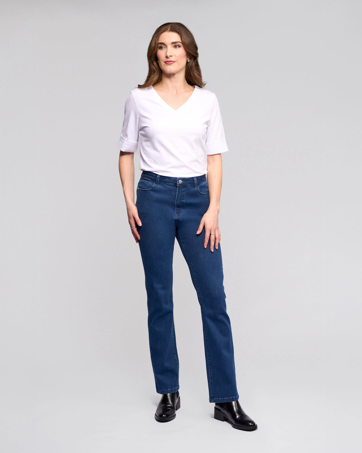 A woman with brown hair and a neutral expression stands against a light gray background, wearing Vassalli's Radio Jean in blue denim, a white V-neck T-shirt, and black ankle boots, with her arms relaxed by her sides.