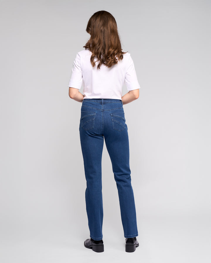 A woman with wavy brown hair faces away, wearing a white short-sleeve shirt, black shoes, and Vassalli's Radio Jean in Blue Denim against a plain light gray background.