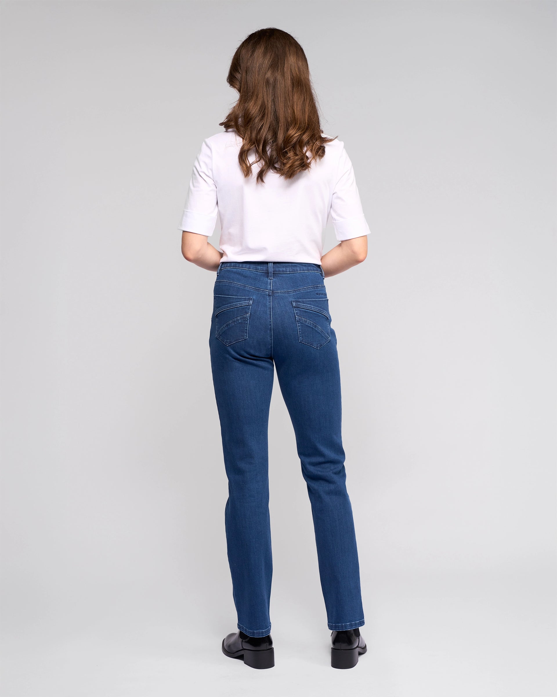 A person with long brown hair faces away, wearing a white short-sleeve shirt, Vassalli Radio Jean - Blue Denim, and black shoes against a plain light gray background.