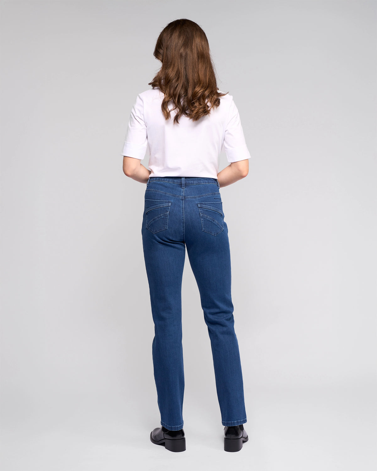 A person with long brown hair faces away, wearing a white short-sleeve shirt, Vassalli Radio Jean - Blue Denim, and black shoes against a plain light gray background.