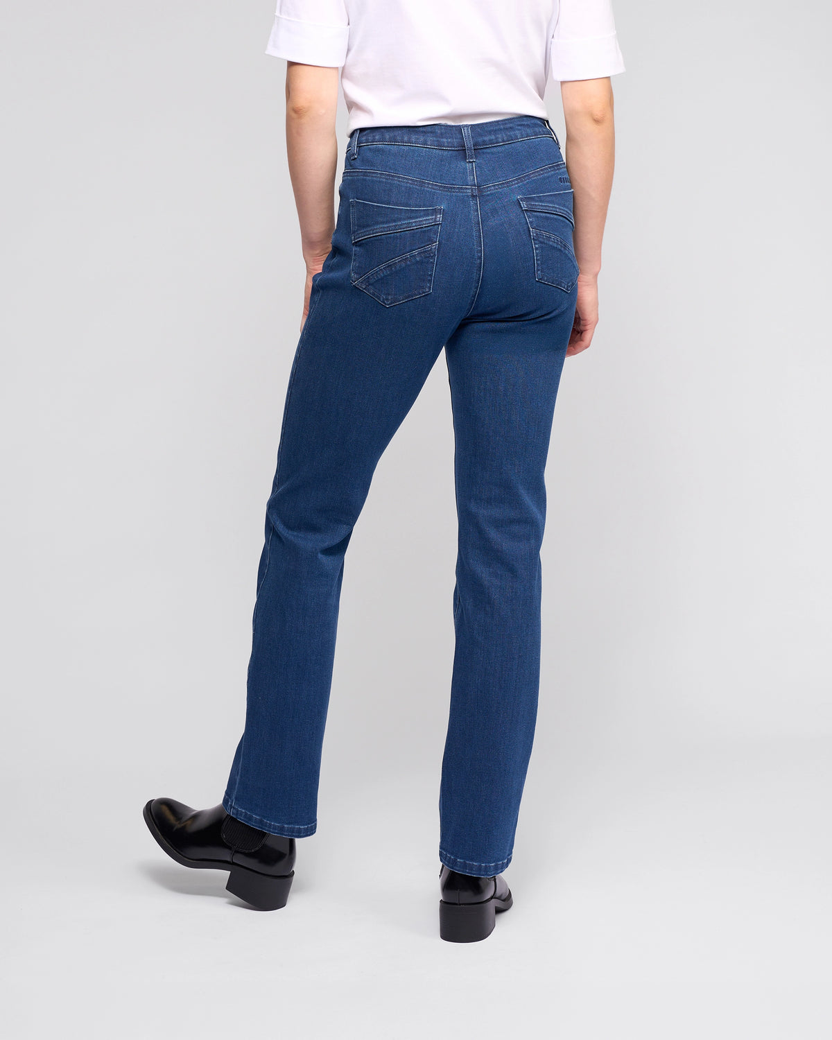 A person is seen from the back, wearing Vassalli's Radio Jean - Blue Denim with visible back pocket stitching, paired with a white short-sleeve shirt and black shoes, standing against a plain background.