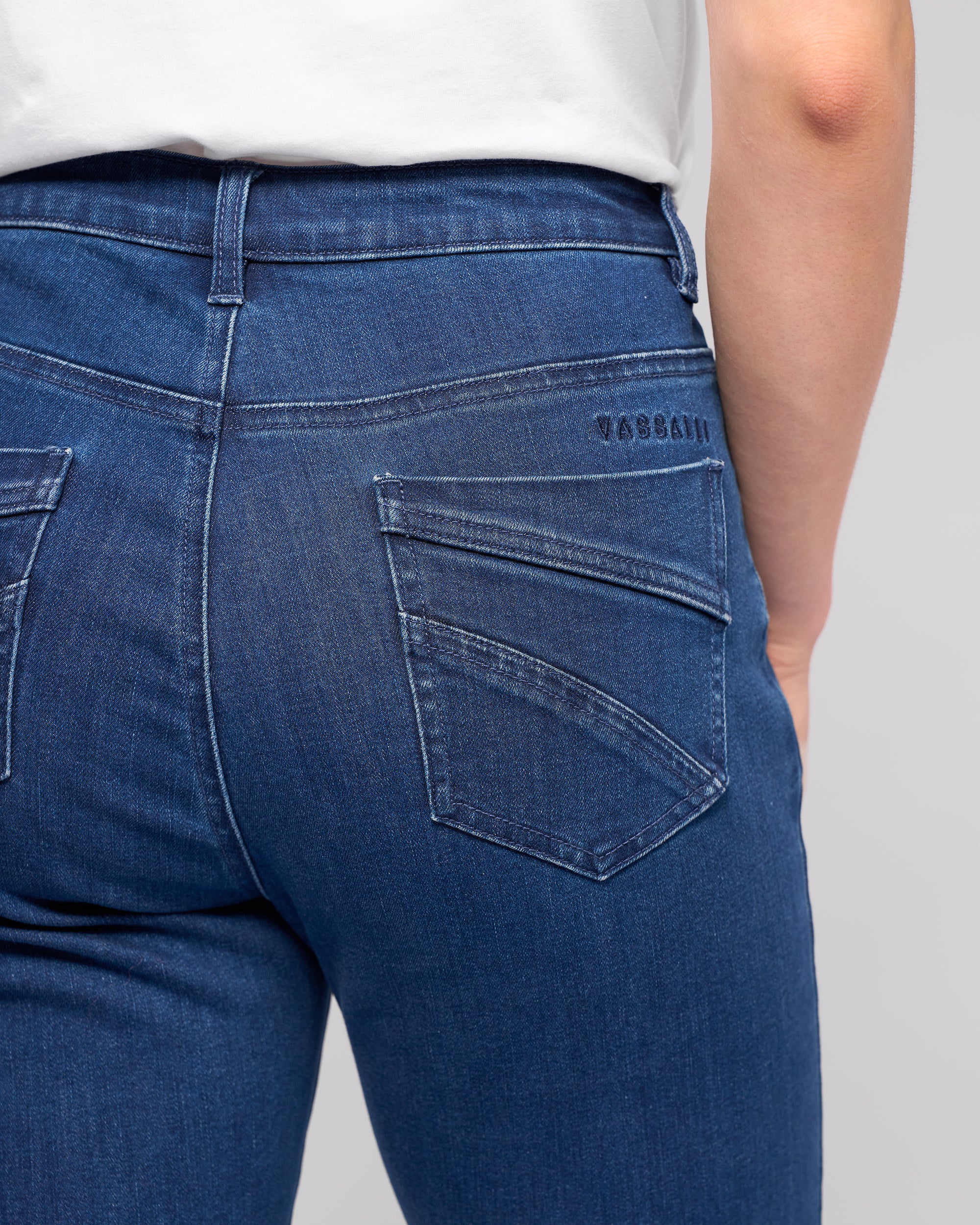 A close-up shows someone wearing Vassalli’s Radio Jean in blue denim with a white shirt, focusing on the back pocket and waist area of the jeans.