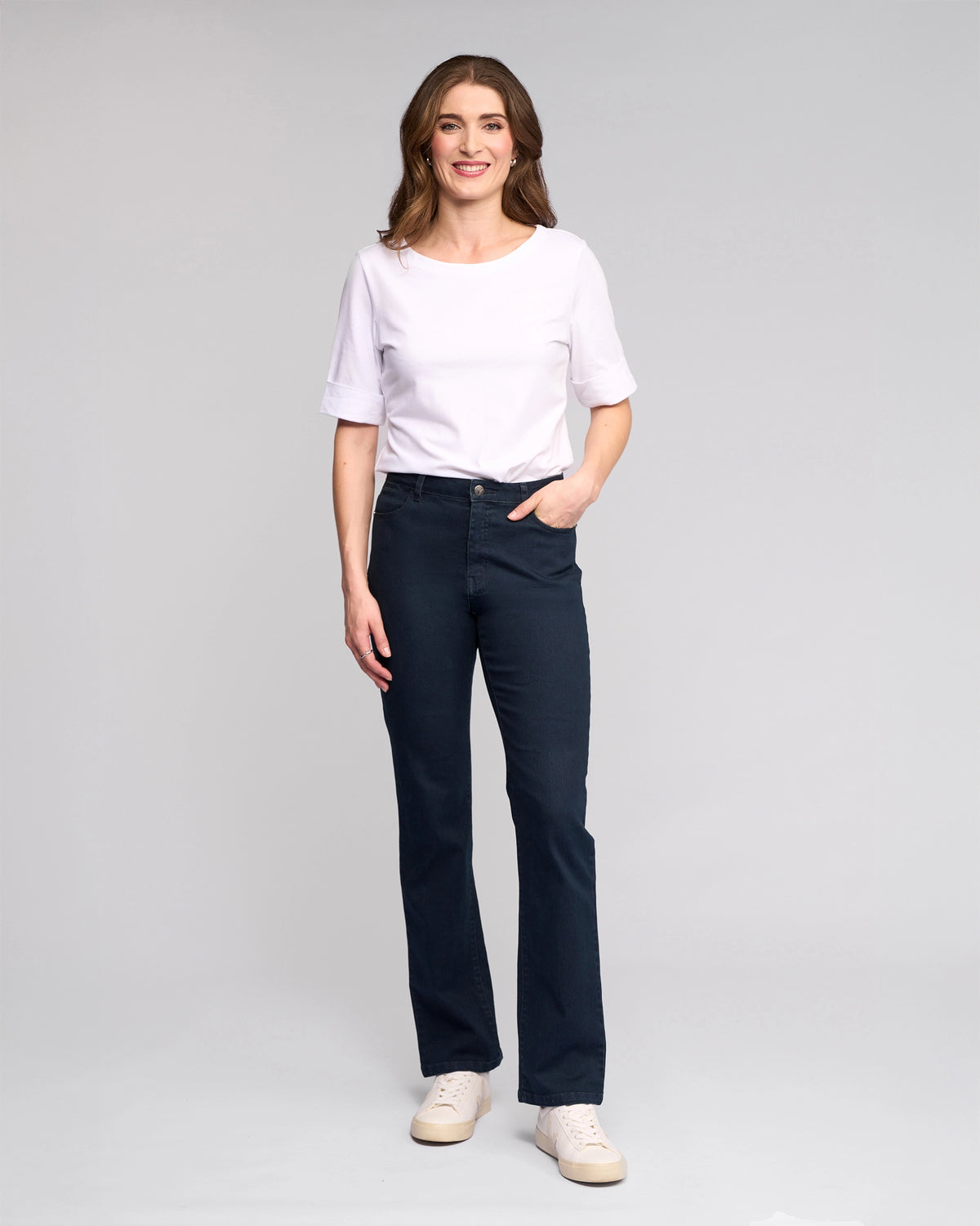 A woman with brown hair smiles in a white short-sleeve shirt, Vassalli's Radio Jean in Indigo Denim, and white sneakers. She stands with one hand in her pocket against a plain light gray background.