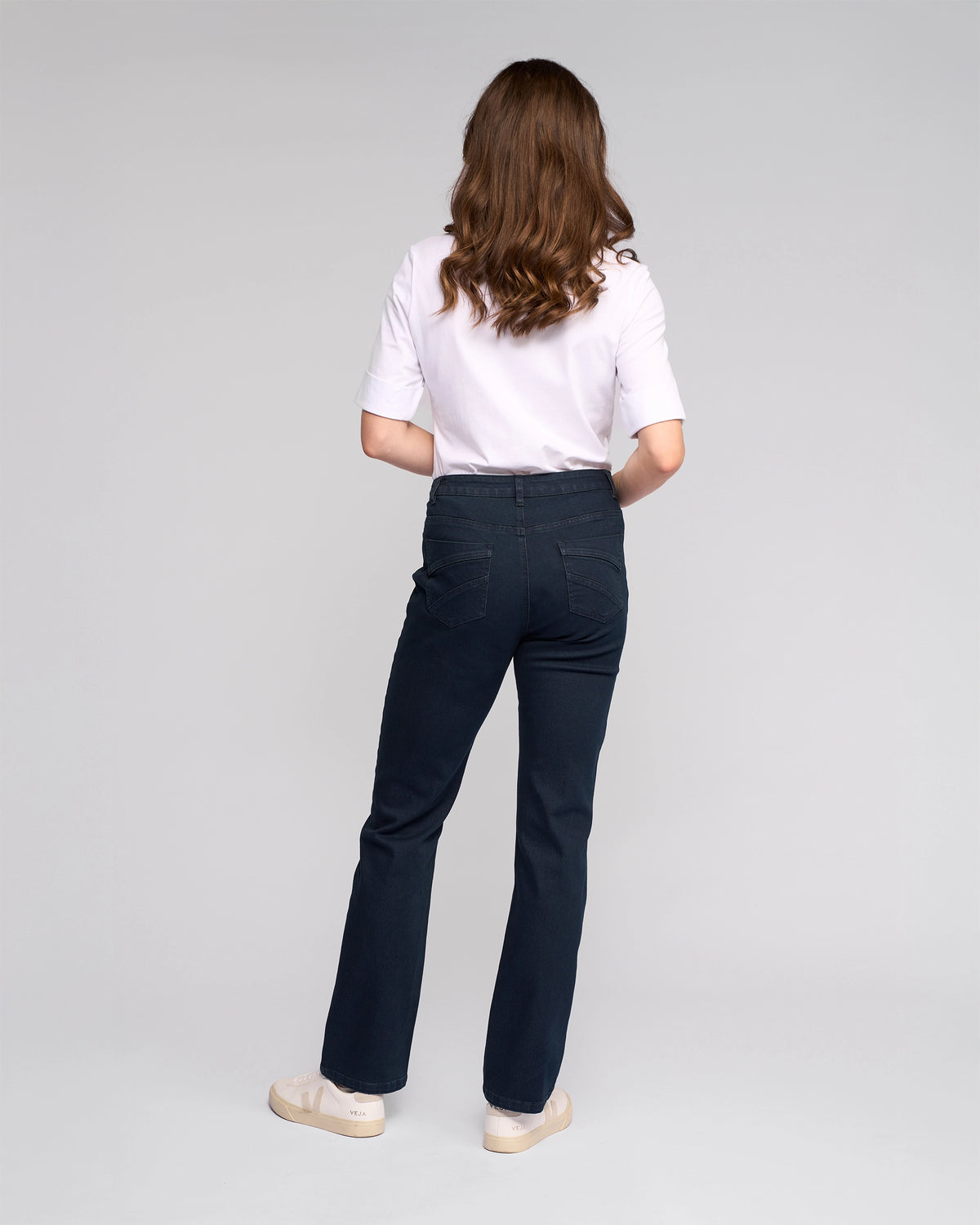 A person with wavy brown hair stands facing away, wearing a white short-sleeve top, beige shoes, and Vassalli’s Radio Jean in Indigo Denim against a plain light gray background.