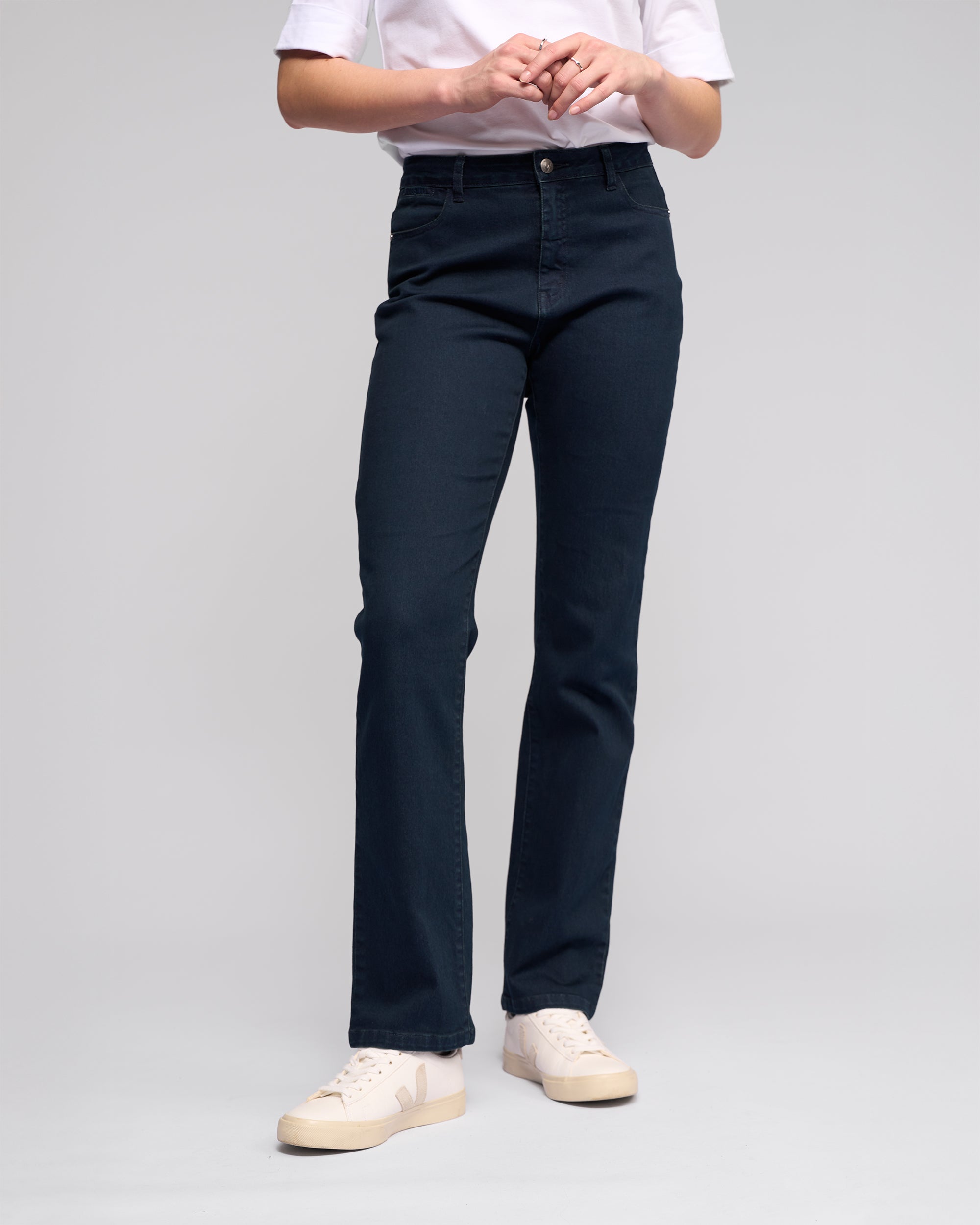Person wearing a white t-shirt, Vassalli Radio Jean in indigo denim, and white sneakers, standing against a plain light background with hands clasped at waist.